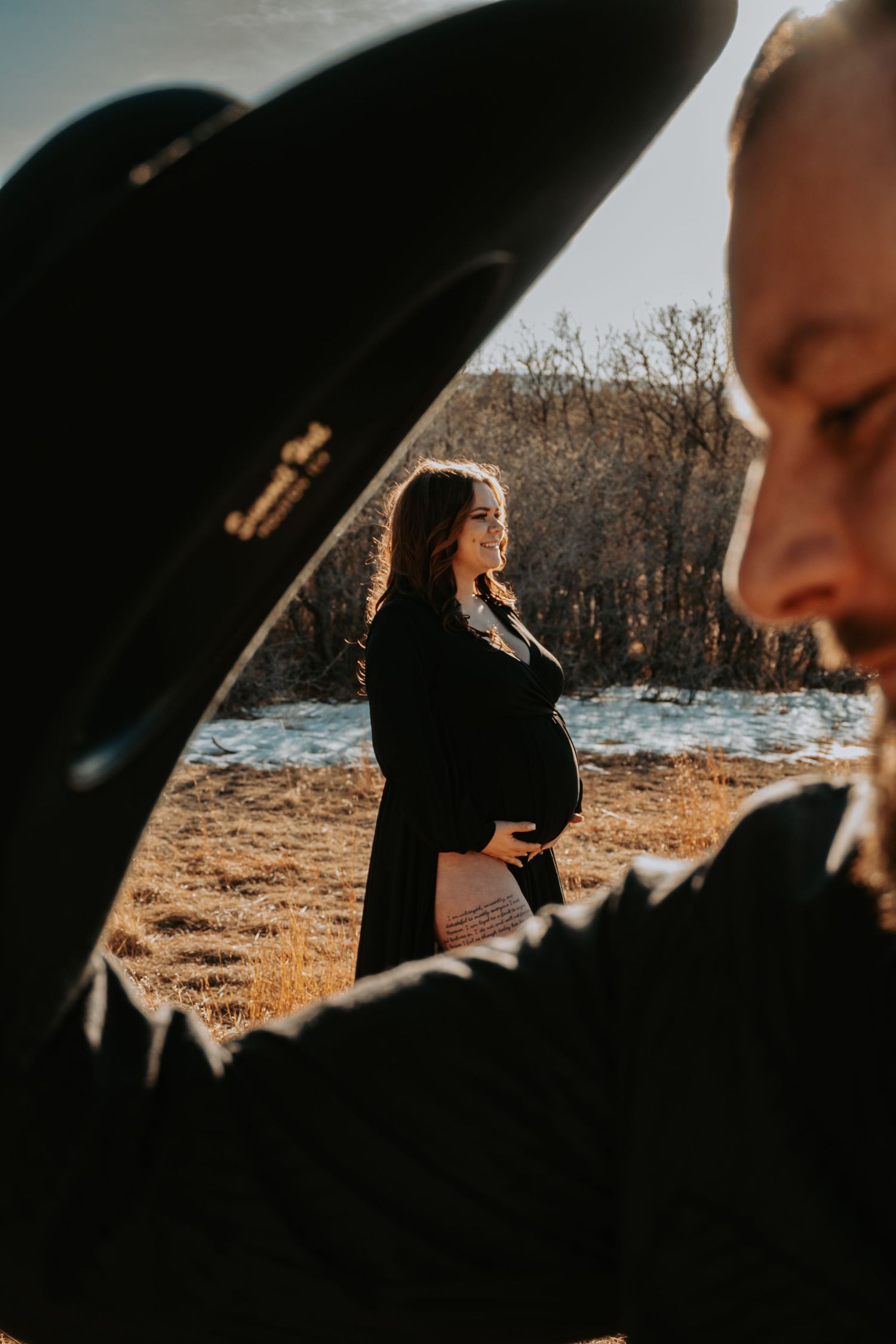 Pregnant woman in black maternity robe smiles while cradling bump in sunny winter field setting.