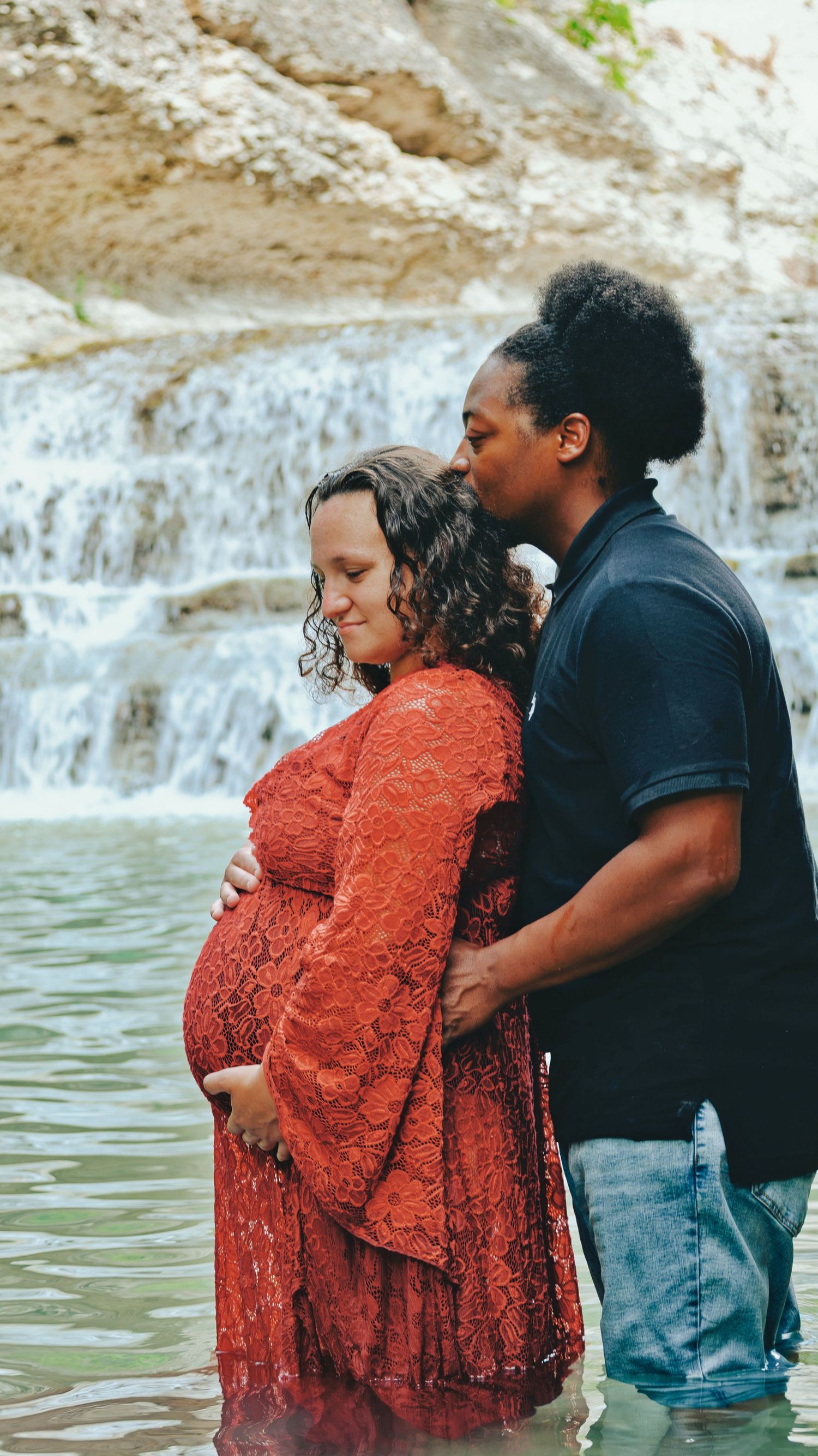 Man kisses pregnant partner tenderly while standing in water before a scenic waterfall backdrop.