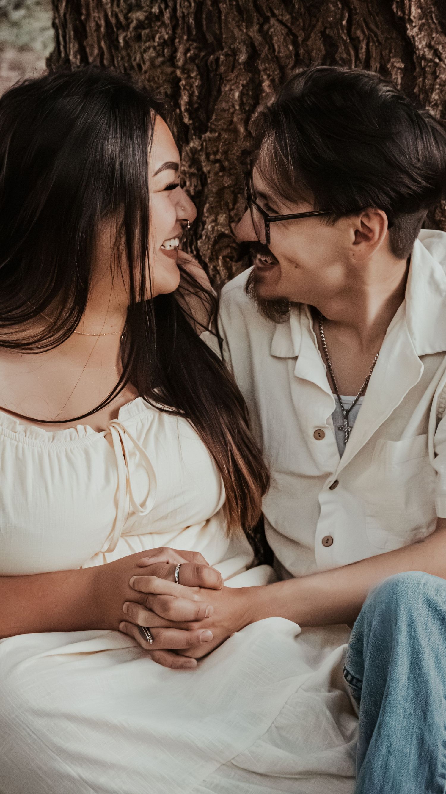 A couple holds hands and smiles at each other while leaning against a tree.