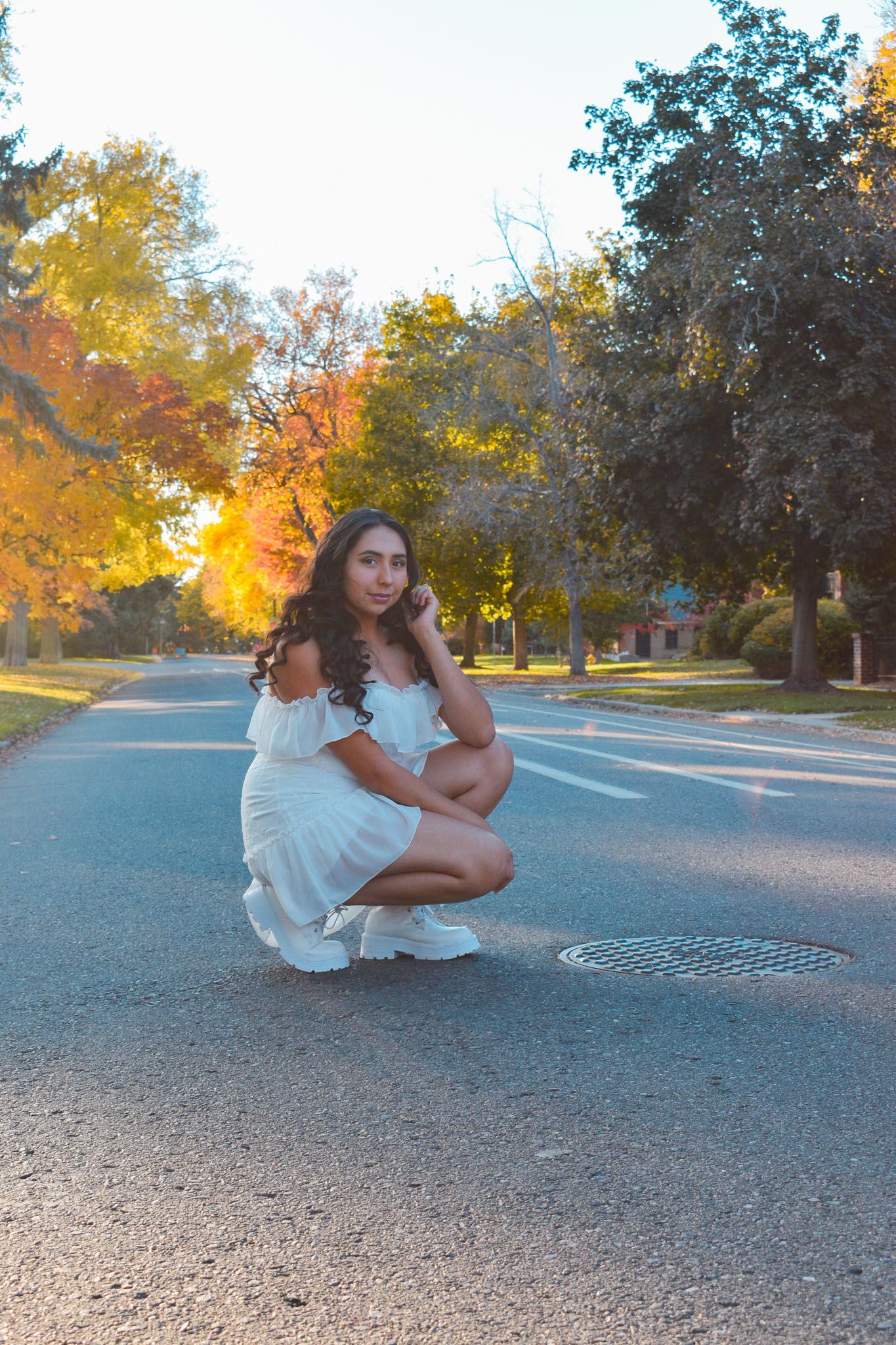 Woman in white ruffled dress crouching on sunlit road surrounded by colorful fall trees for senior photos.