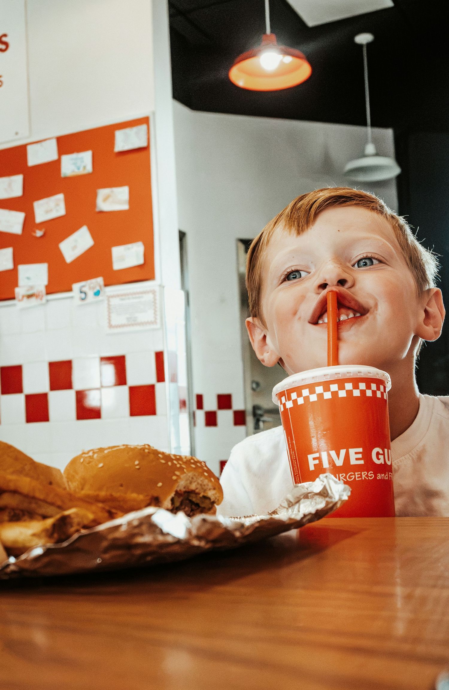 Young boy drinking from a Five Guys cup with a straw, burger and fries on table before him.