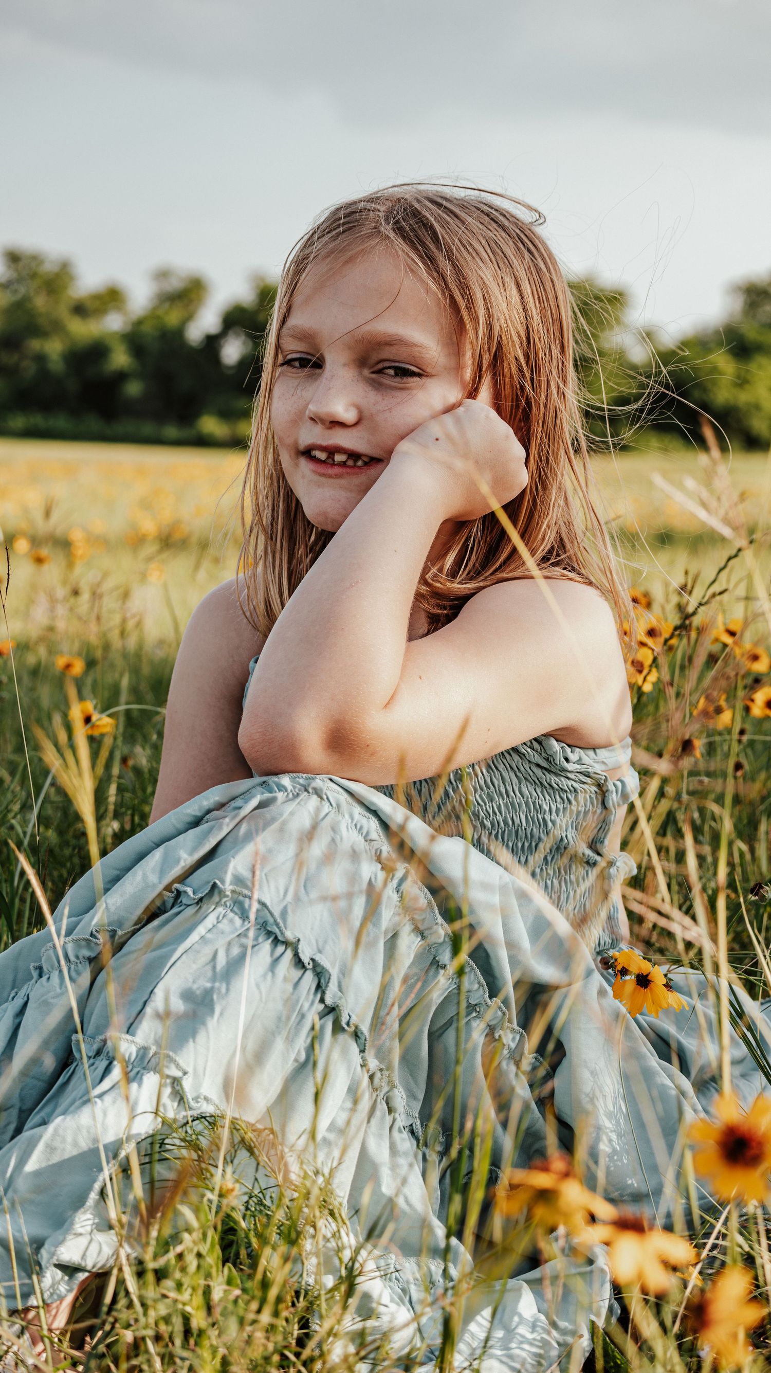 Smiling young girl in light blue dress resting chin on hand in sunny wildflower field.