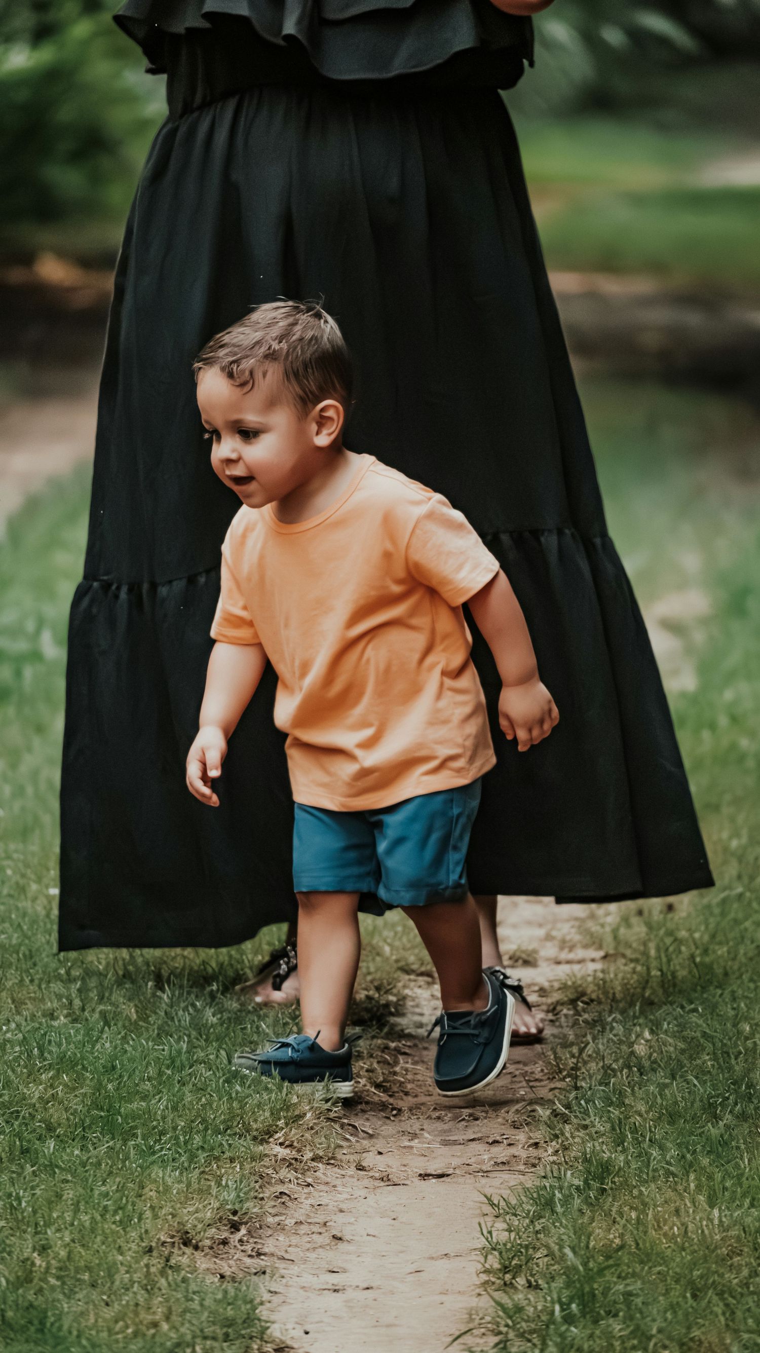 Toddler boy in orange shirt and blue shorts walking on a garden path.