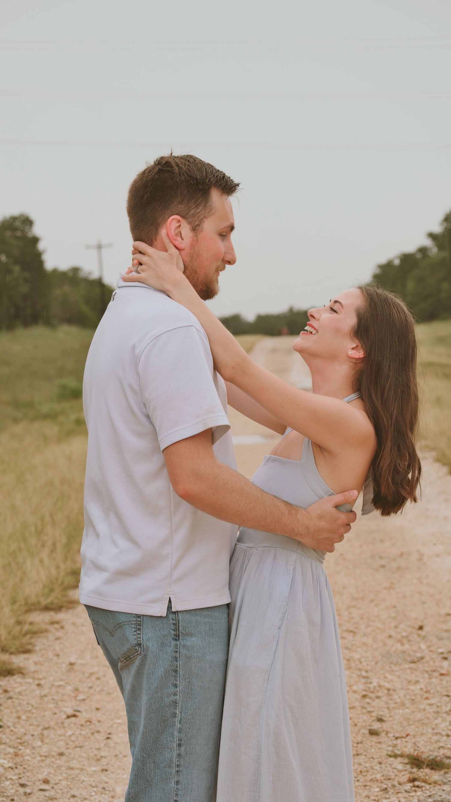 A smiling couple embraces on a rural dirt road, laughing joyfully at each other for a family session.