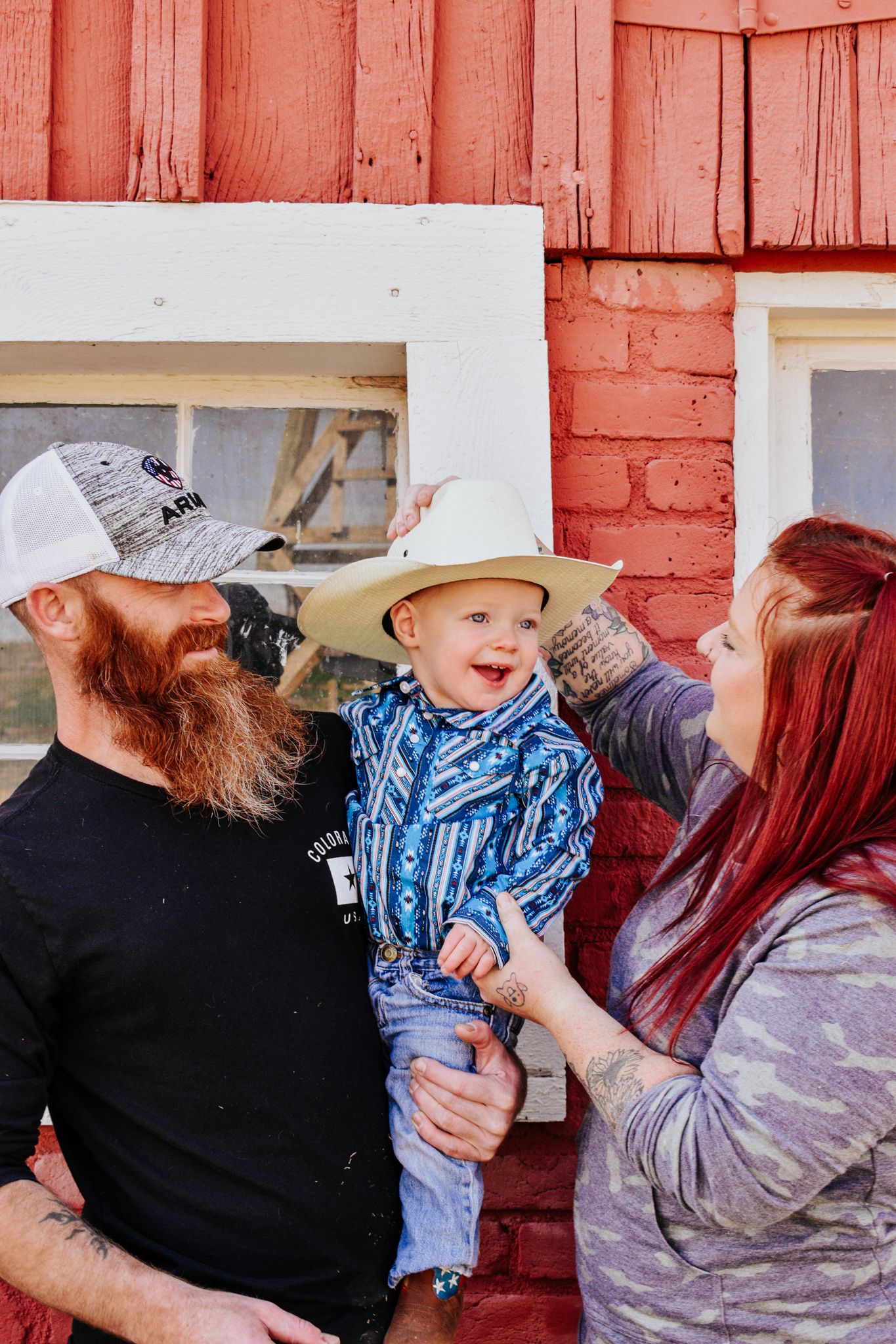 Parents adjusting cowboy hat on laughing baby boy outside red barn building. Family Photographer in Pflugerville, Tx