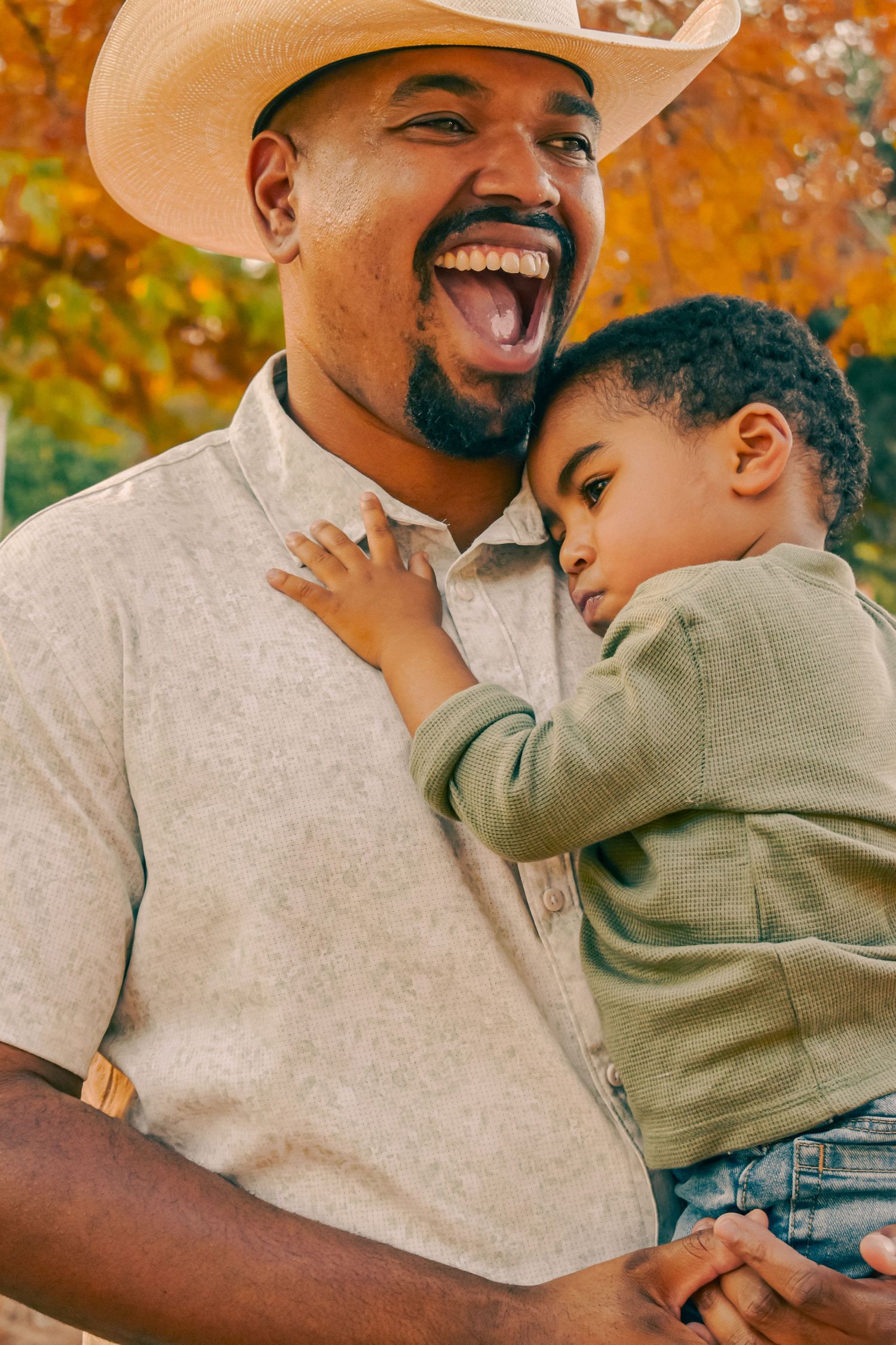 A joyful father in a straw cowboy hat laughs while cradling his sleepy toddler in an autumn park.