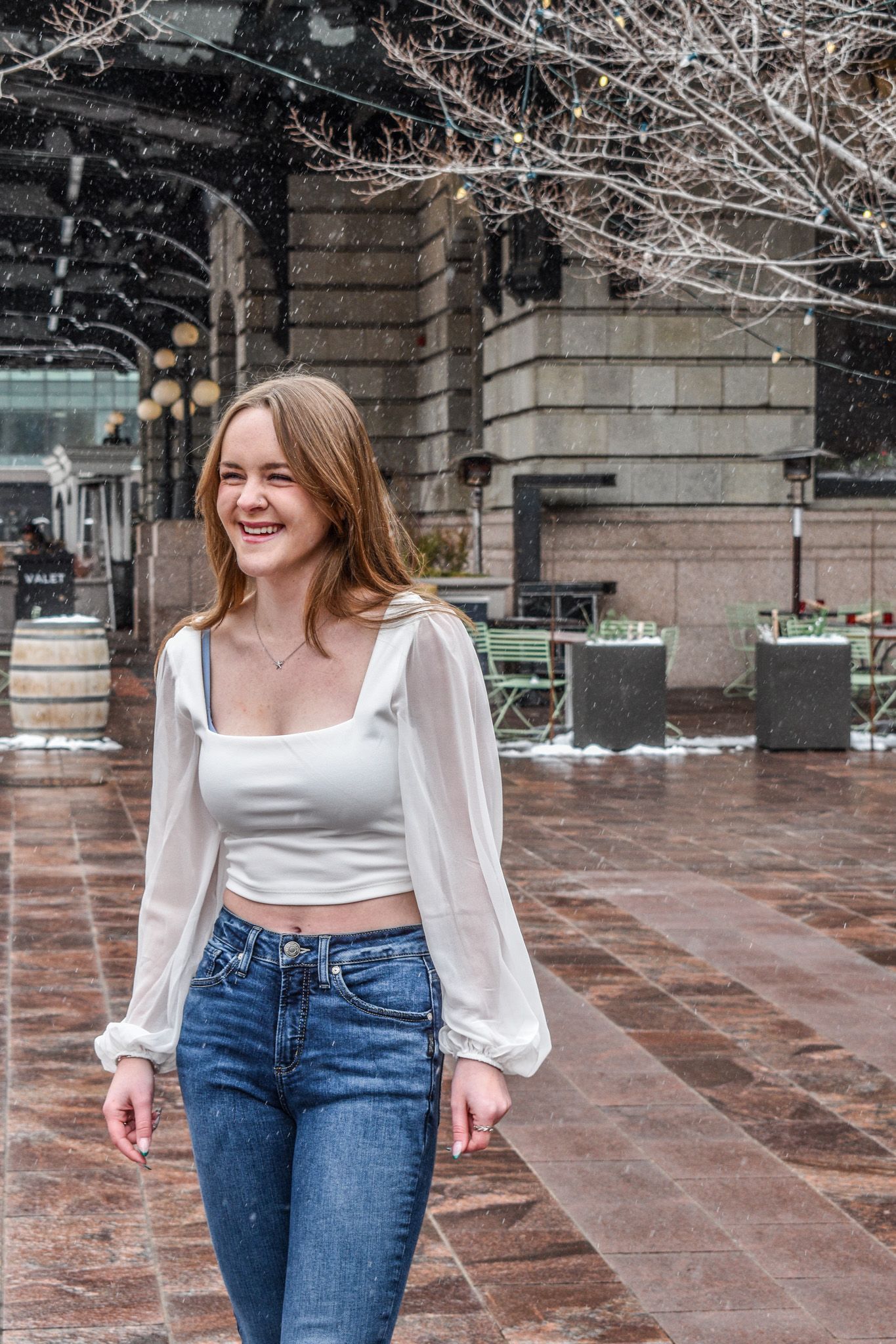 Laughing young woman in white crop top and blue jeans walking through snowy city plaza in winter for senior photos.