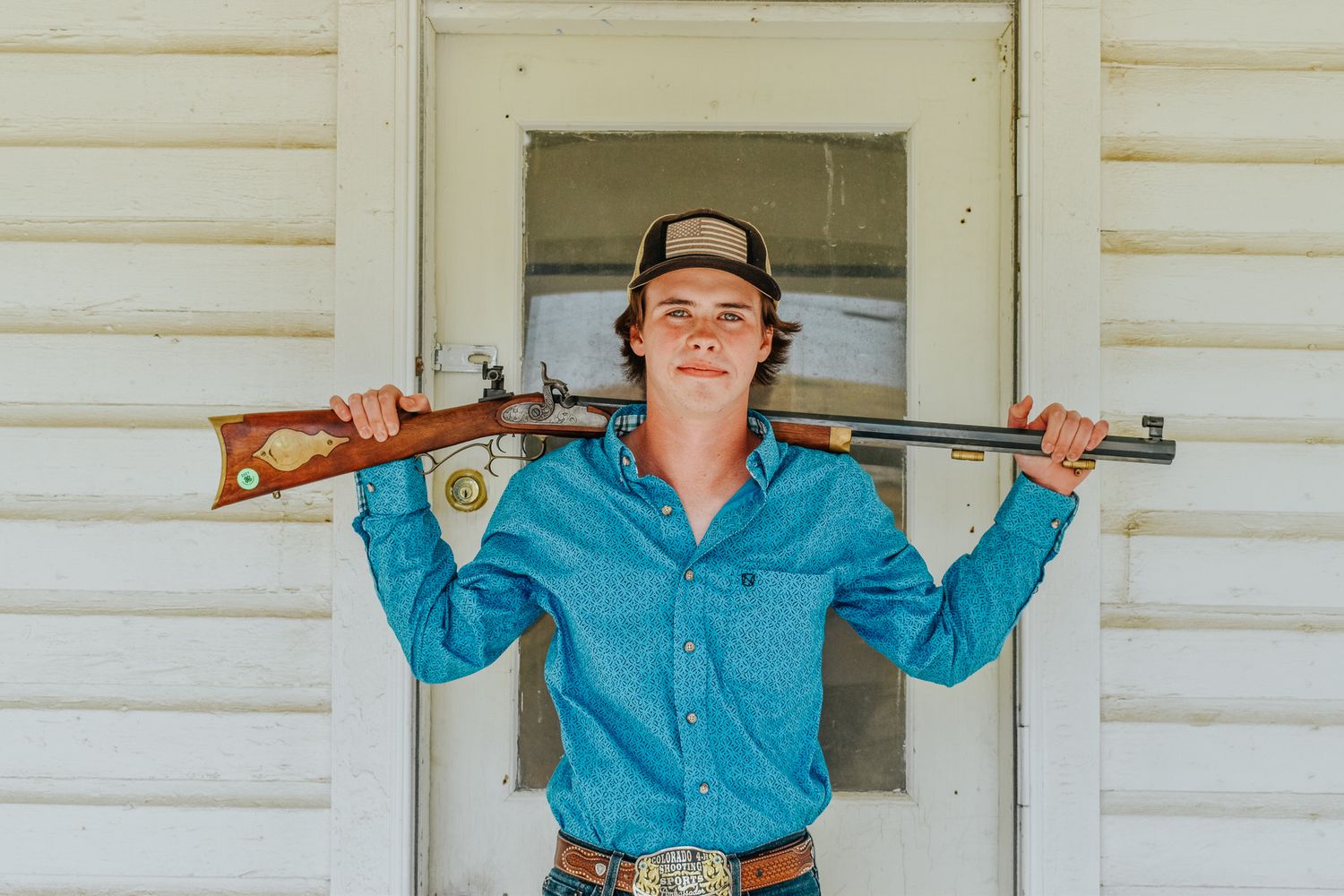 Young man in blue western shirt holding a rifle across his shoulders outside a white building for senior photos.