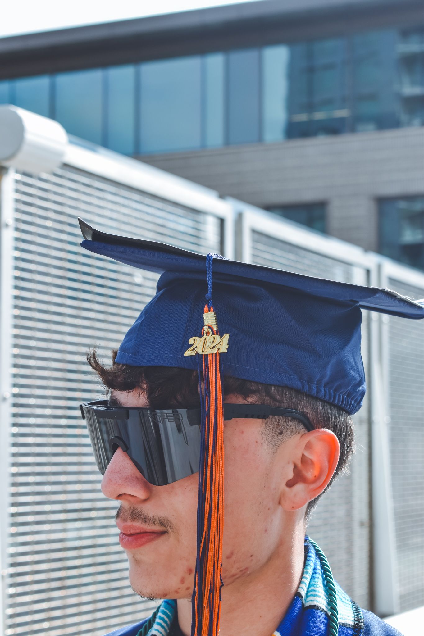 Close-up of graduate wearing blue 2024 mortarboard cap with orange tassel and dark sunglasses for senior photos.
