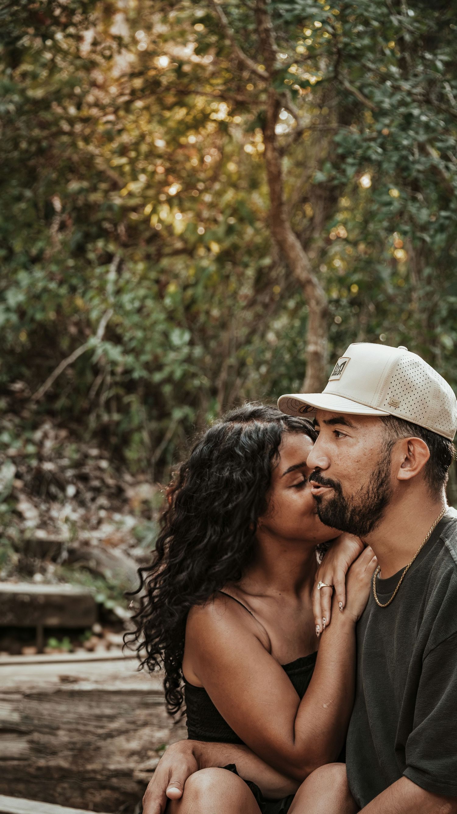Couple sharing a kiss outdoors in a lush green forest setting, woman wearing an engagement ring.