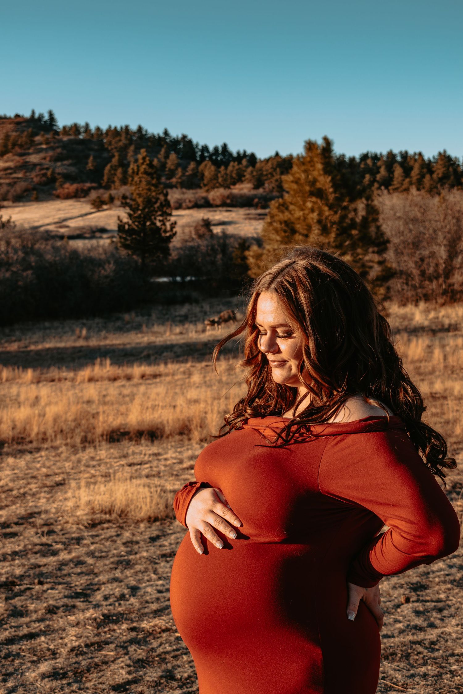 Pregnant woman in red off-shoulder dress gently holds baby bump in autumn landscape.