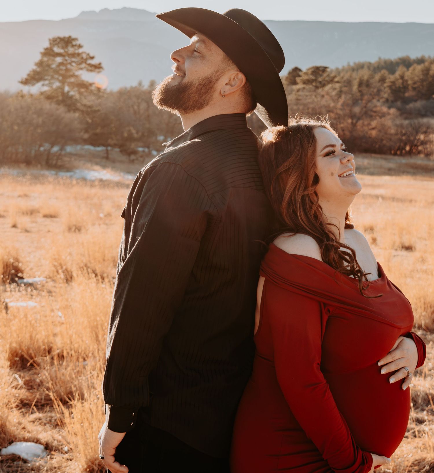 Joyful maternity couple standing back-to-back laughing in golden meadow at sunset.