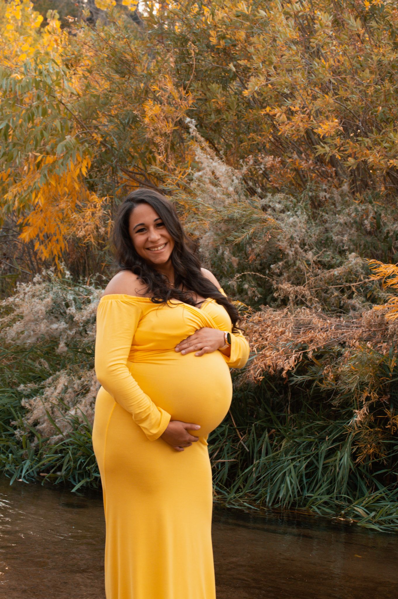 Smiling pregnant woman in yellow off-shoulder maternity gown standing near creek with vibrant autumn foliage backdrop.