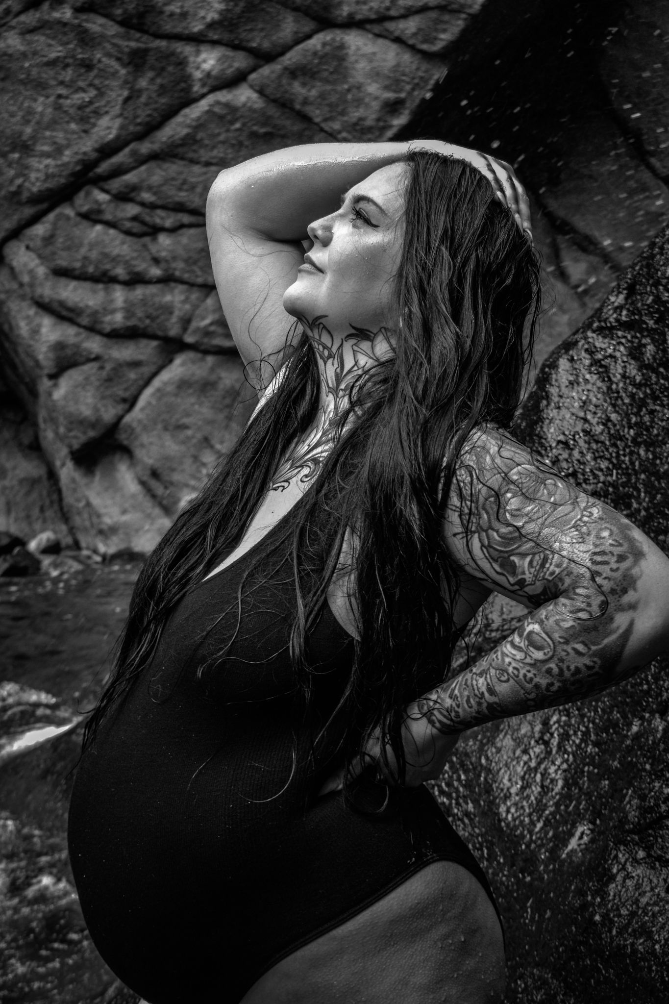 Black and white portrait of tattooed pregnant woman in swimsuit posing against large rocks.