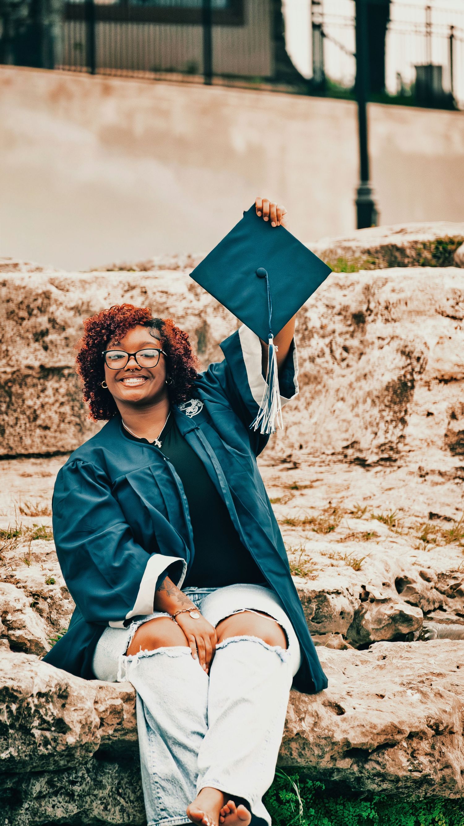 Smiling graduate in blue cap and gown sitting on stone wall holding mortarboard cap outdoors for senior photos.