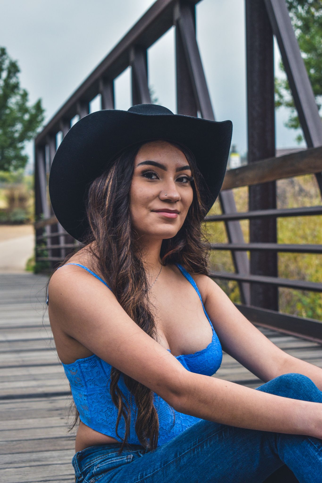 Young woman in black cowboy hat and blue crop top sits on a wooden bridge, smiling softly for senior photos.