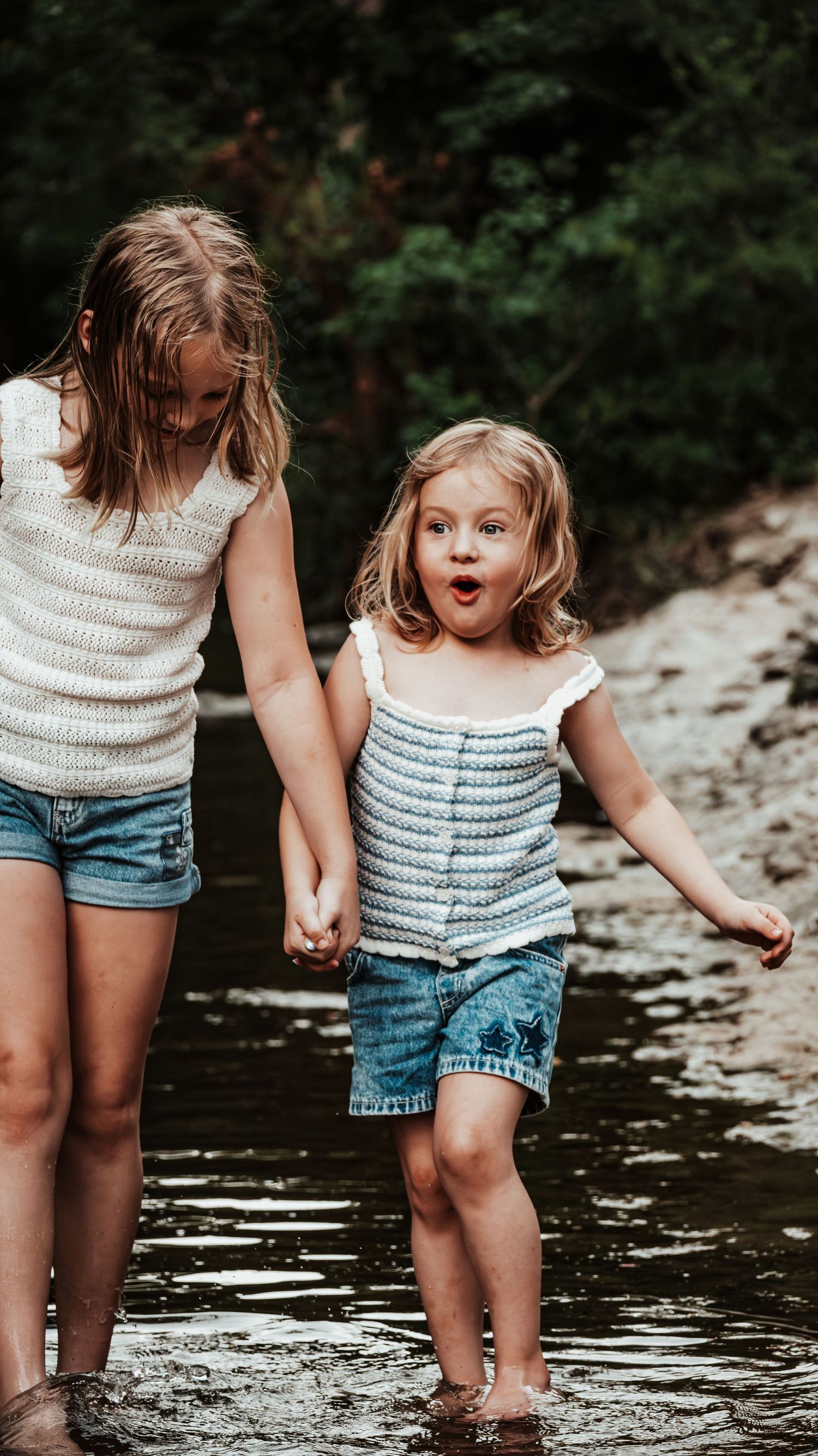 Two young sisters holding hands wading barefoot in a shallow creek surrounded by trees.