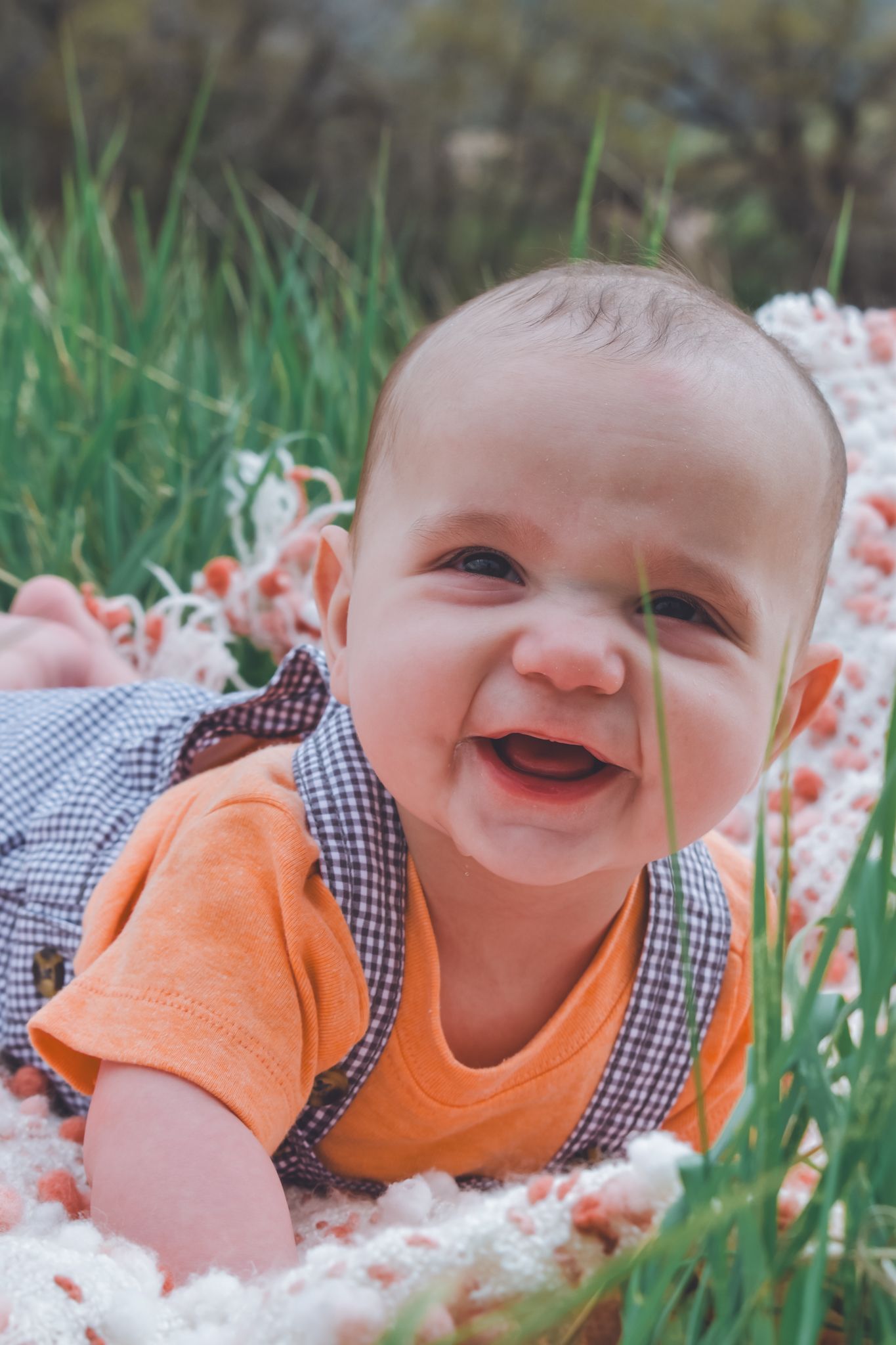 Happy smiling baby in orange shirt lying on white blanket in green grass outdoors. Family Photographer in Pflugerville, Tx