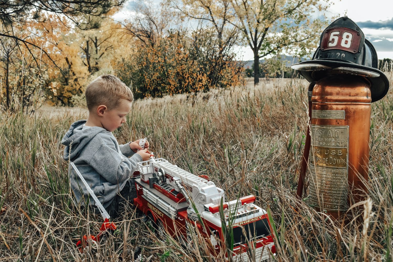 Young boy sits in tall autumn grass playing with toy fire truck next to vintage fire extinguisher. Family Photographer in Pflugerville, Tx