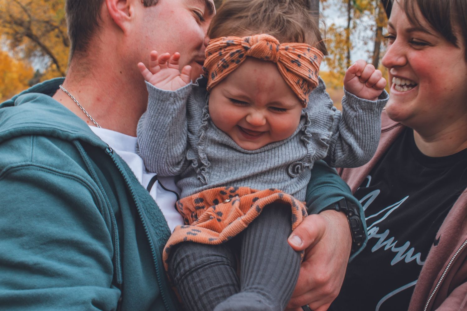 Two parents holding and kissing their smiling baby girl outdoors in autumn. Family Photographer in Pflugerville, Tx