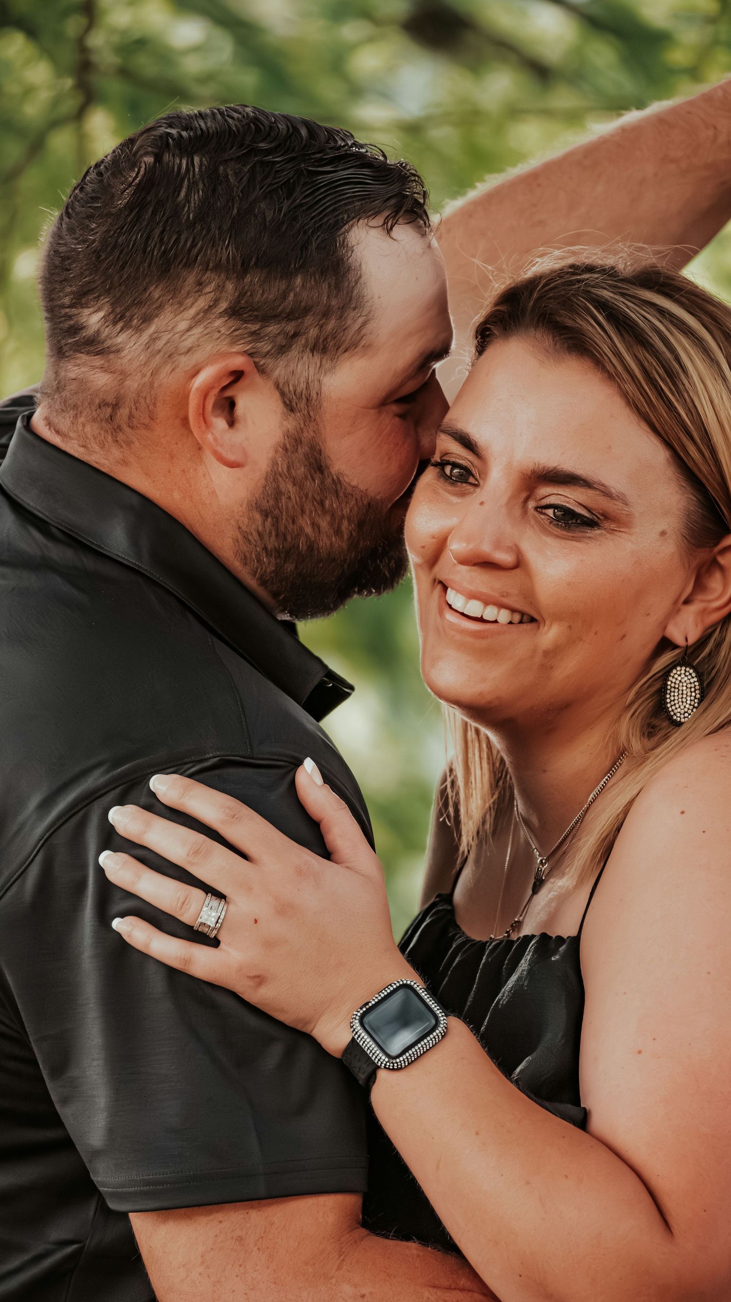 A smiling couple in black outfits share a tender moment outdoors, the man kissing the womans cheek as she beams with joy.
