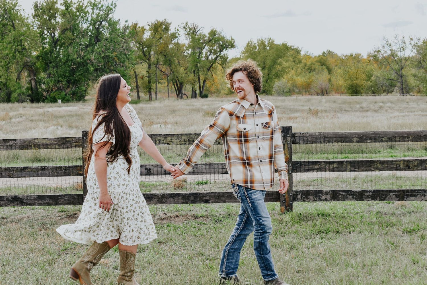 Happy couple walking hand in hand through a farm field with a wooden fence in the background.