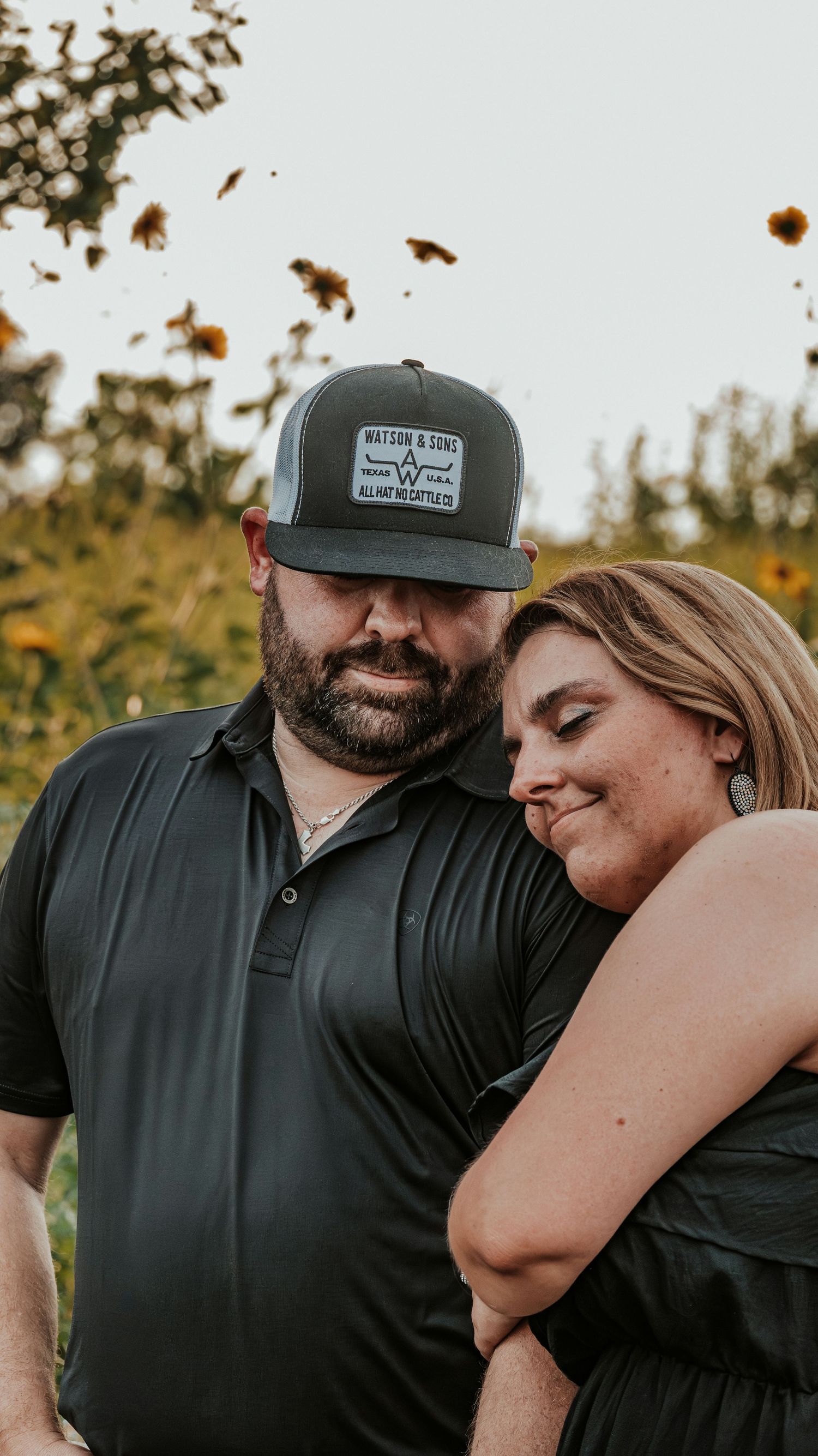 Couple embracing in a sunflower field at dusk, man wearing a Watson and Sons cap.