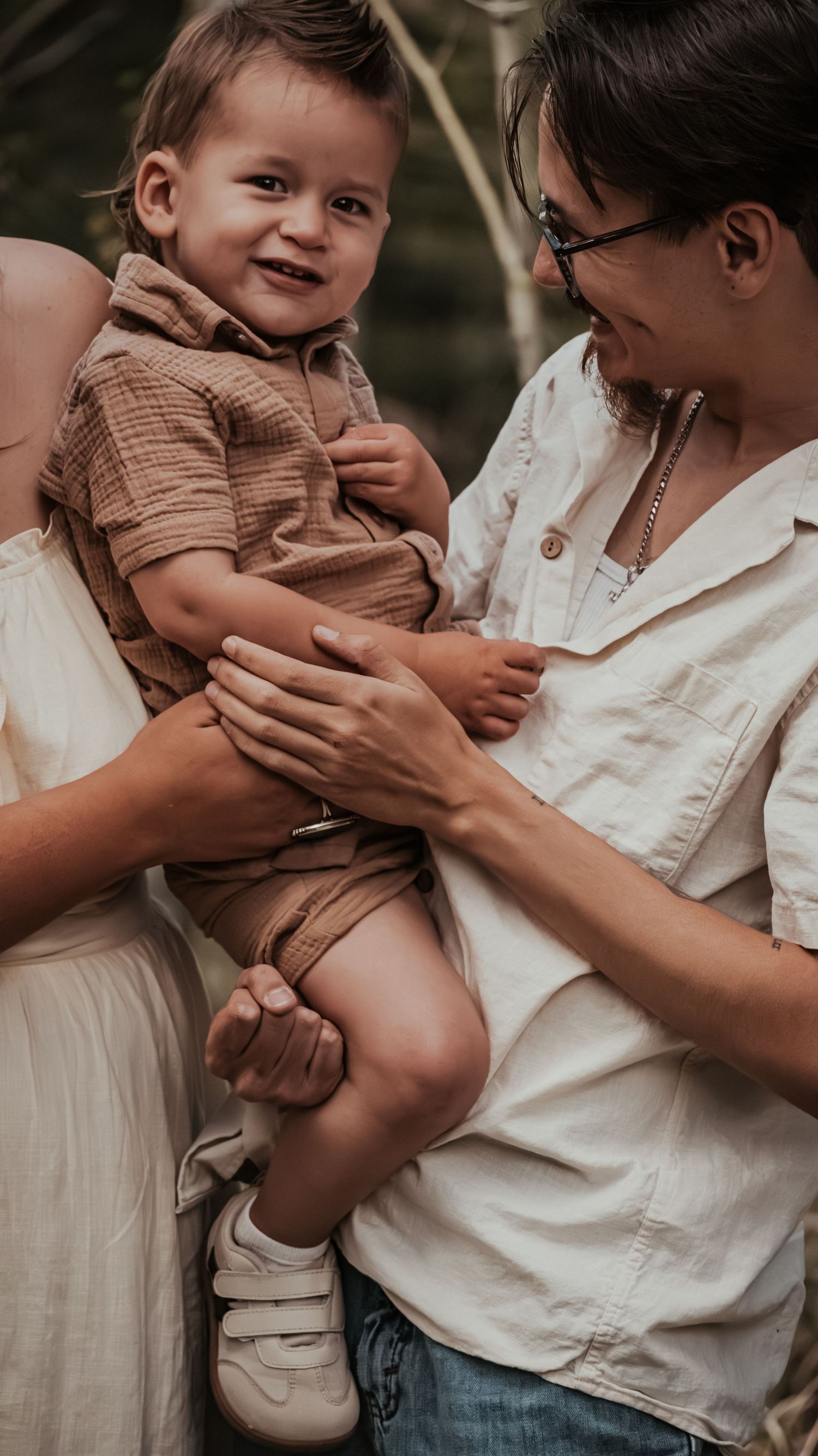 Smiling baby in brown outfit is held by father in white shirt while mother looks on lovingly outdoors.