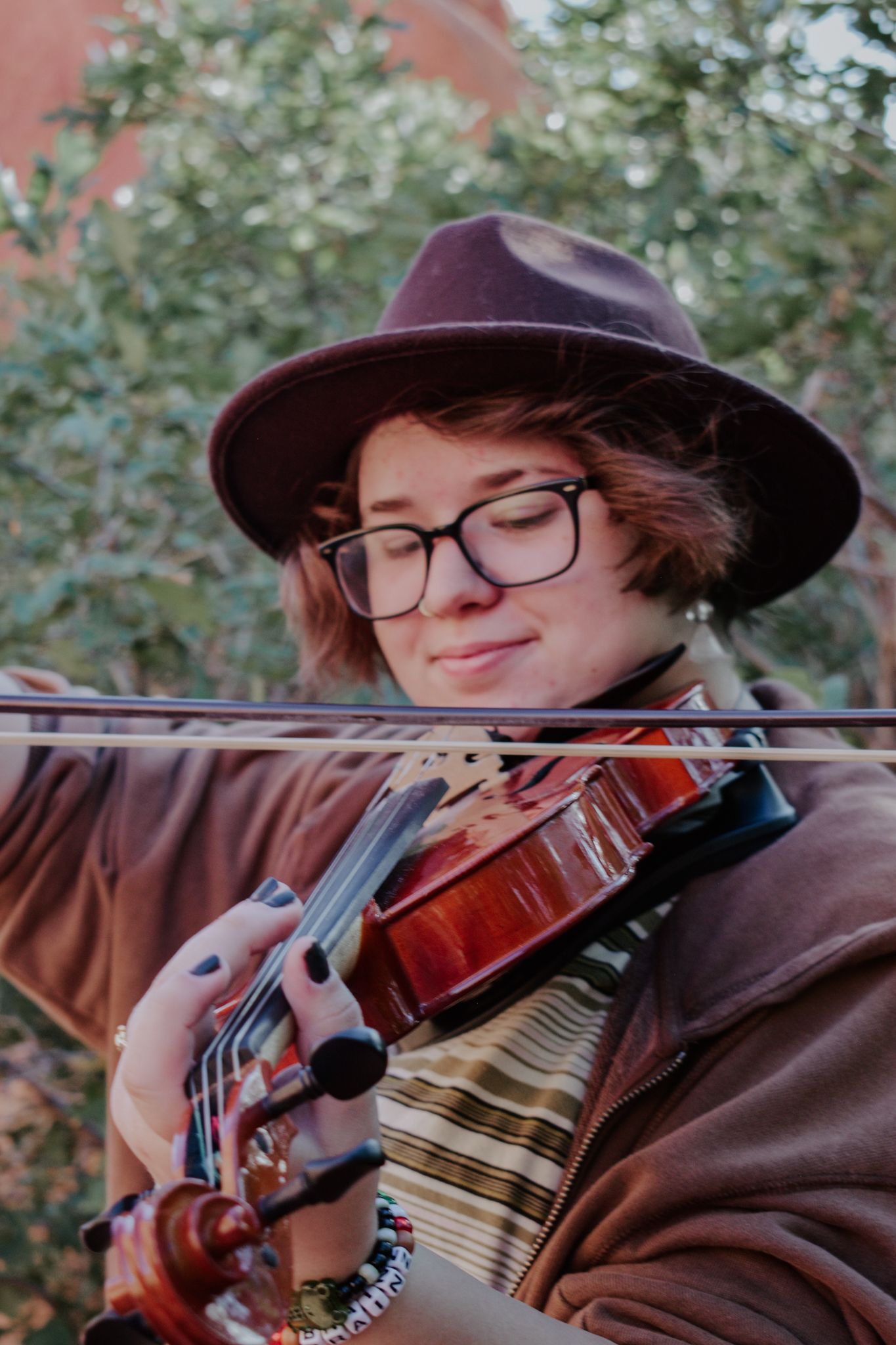 Young musician in brown hat and glasses playing violin outdoors near red rock formations for senior photos.