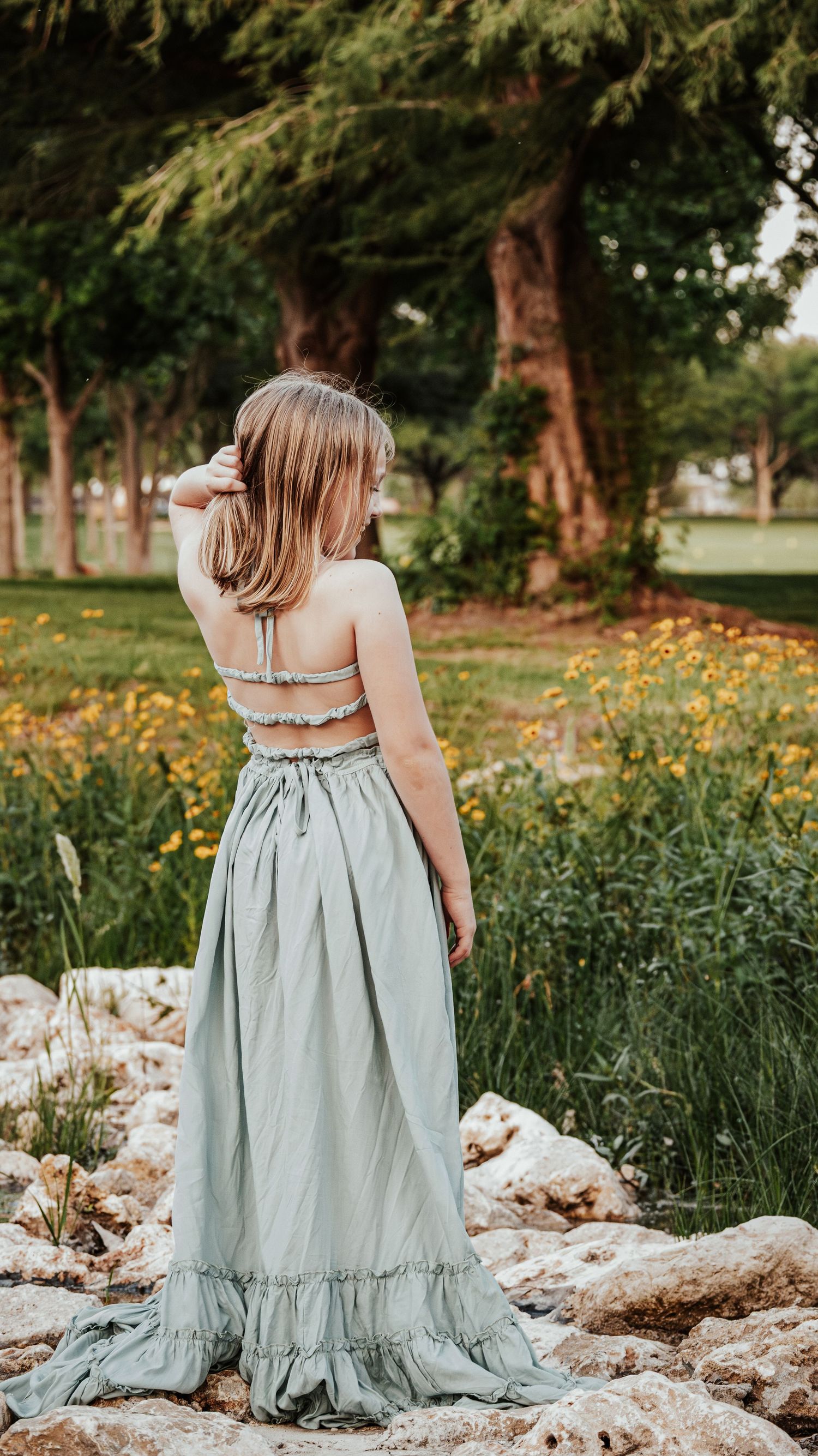 Young girl in sage green maxi dress standing on rocks with yellow wildflowers in background.