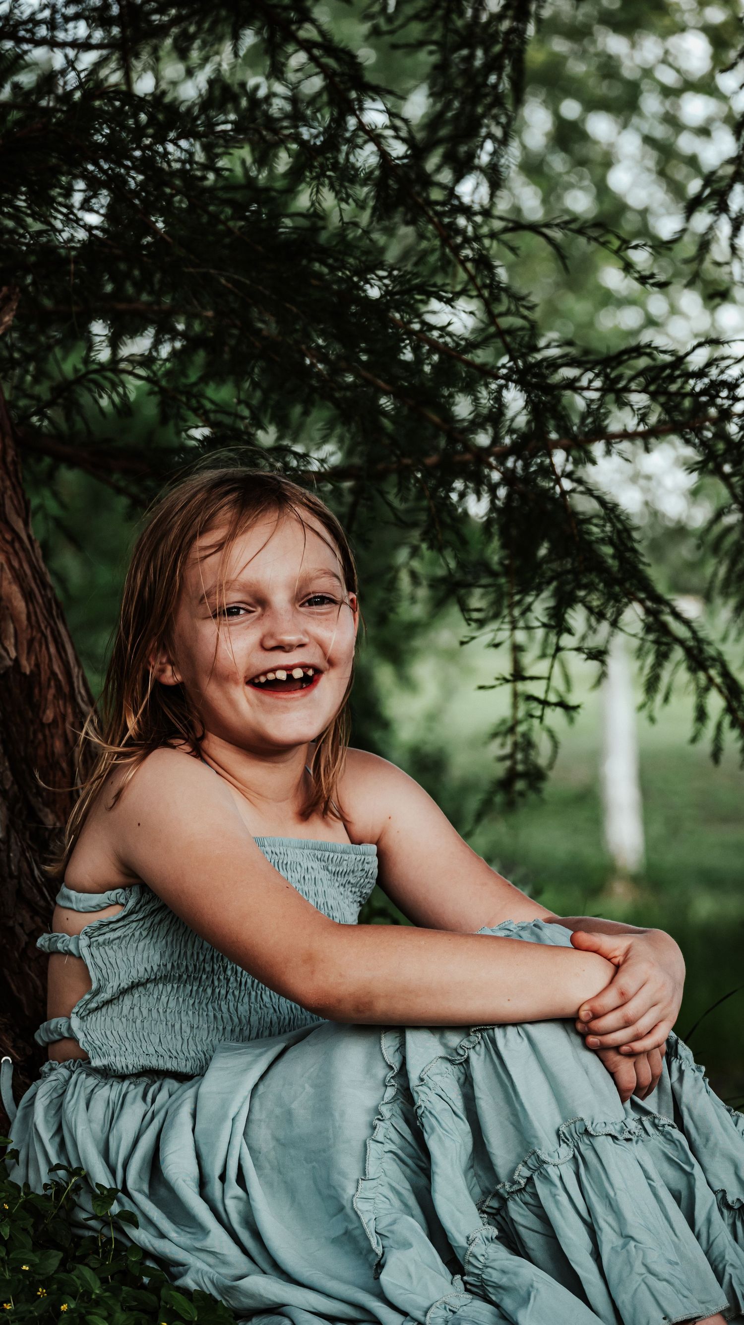 Cheerful young girl in a teal smocked dress laughing while seated near a pine tree outdoors.