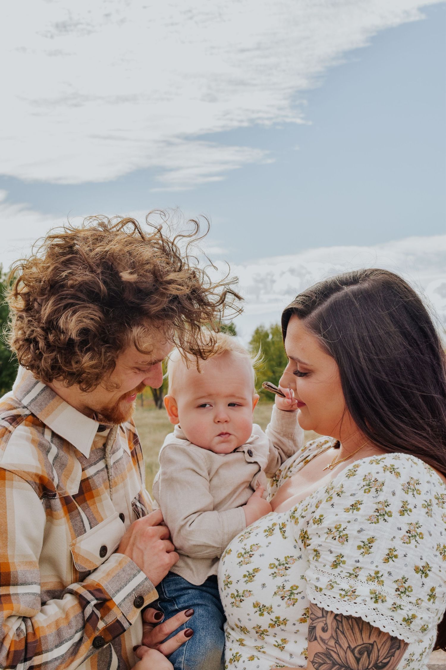 Young family with curly-haired dad and brunette mom holding their baby outdoors.