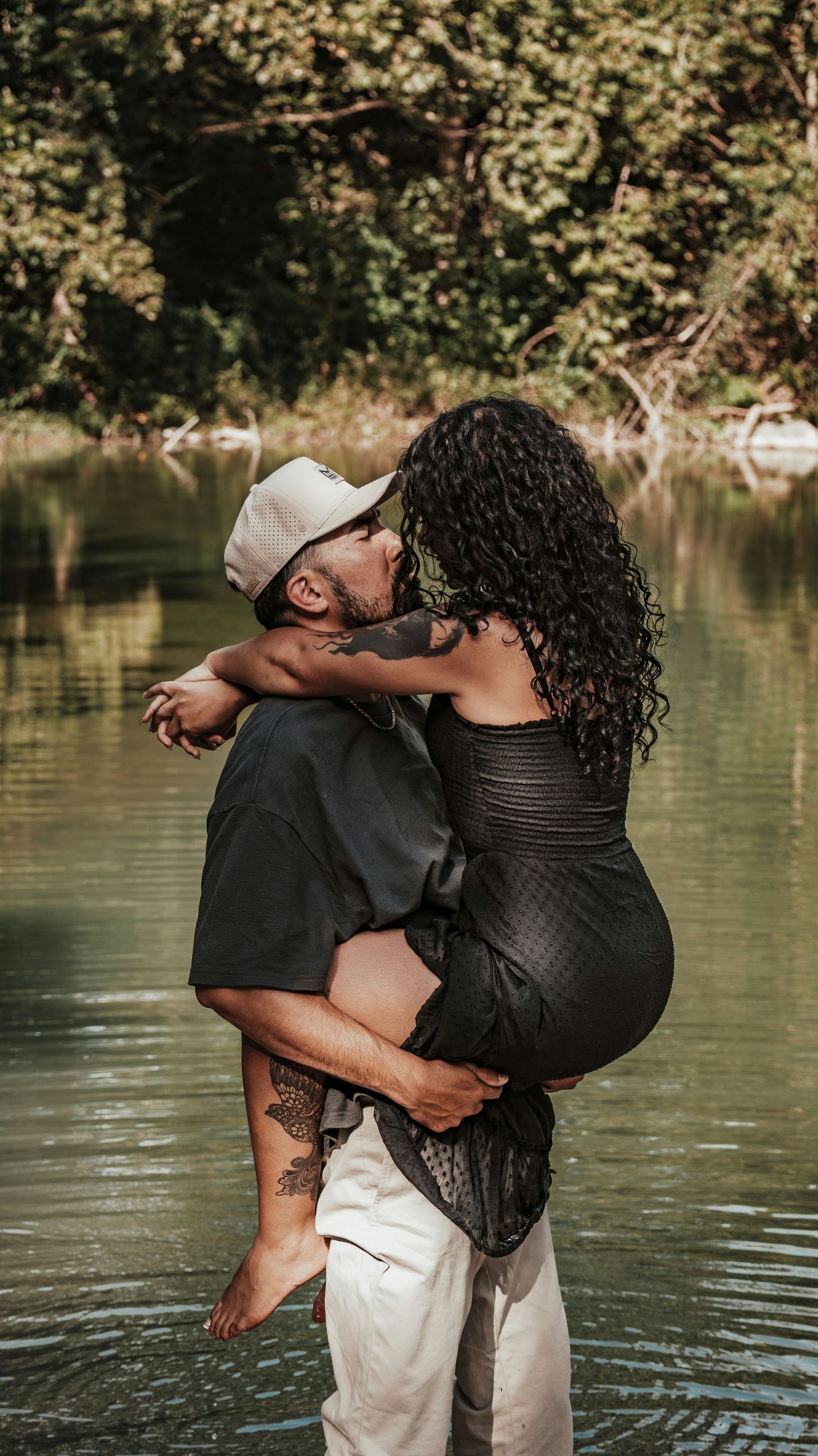 A couple shares a romantic kiss as the man lifts the woman near a lush green river.