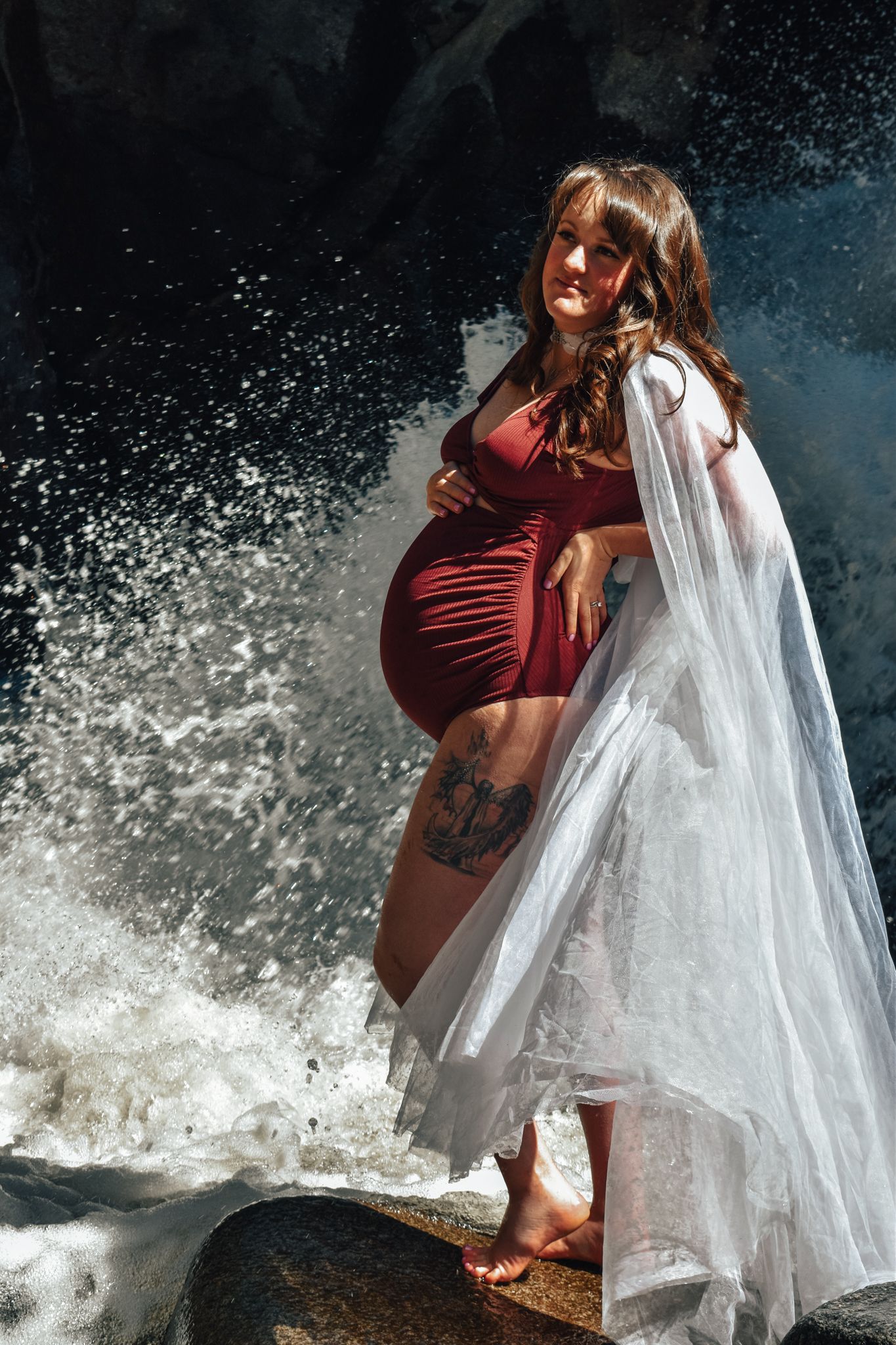 Pregnant woman in red swimsuit and white veil poses by a waterfall with anchor tattoo visible.  Maternity Photographer in Pflugerville, Tx