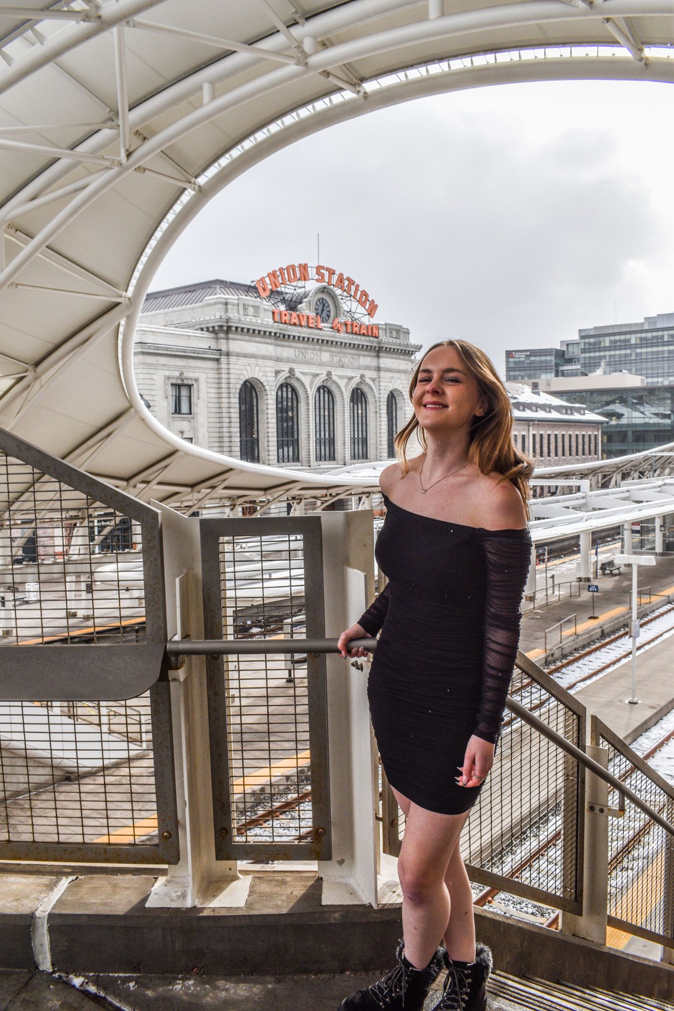 Young woman in black mini dress standing on staircase with Denver Union Station visible in background for senior photos.