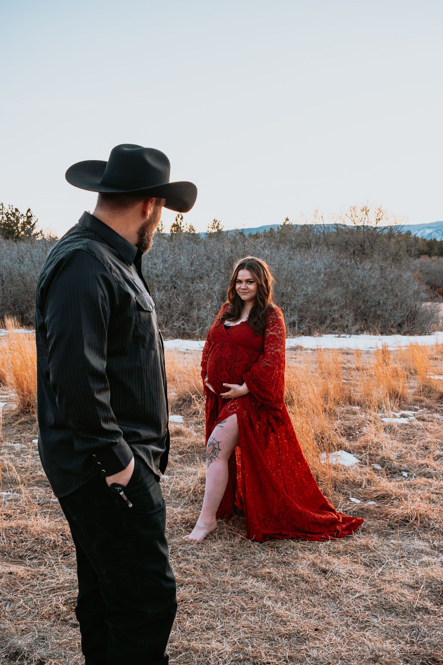 Pregnant woman in red lace gown smiles at partner in black cowboy hat outdoors.