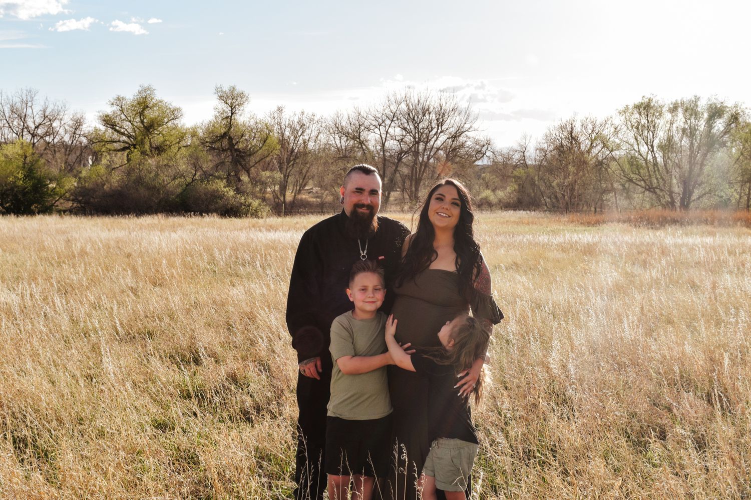 Family of three poses in sunlit golden field during maternity session, father and child flanking pregnant mother in olive dress.