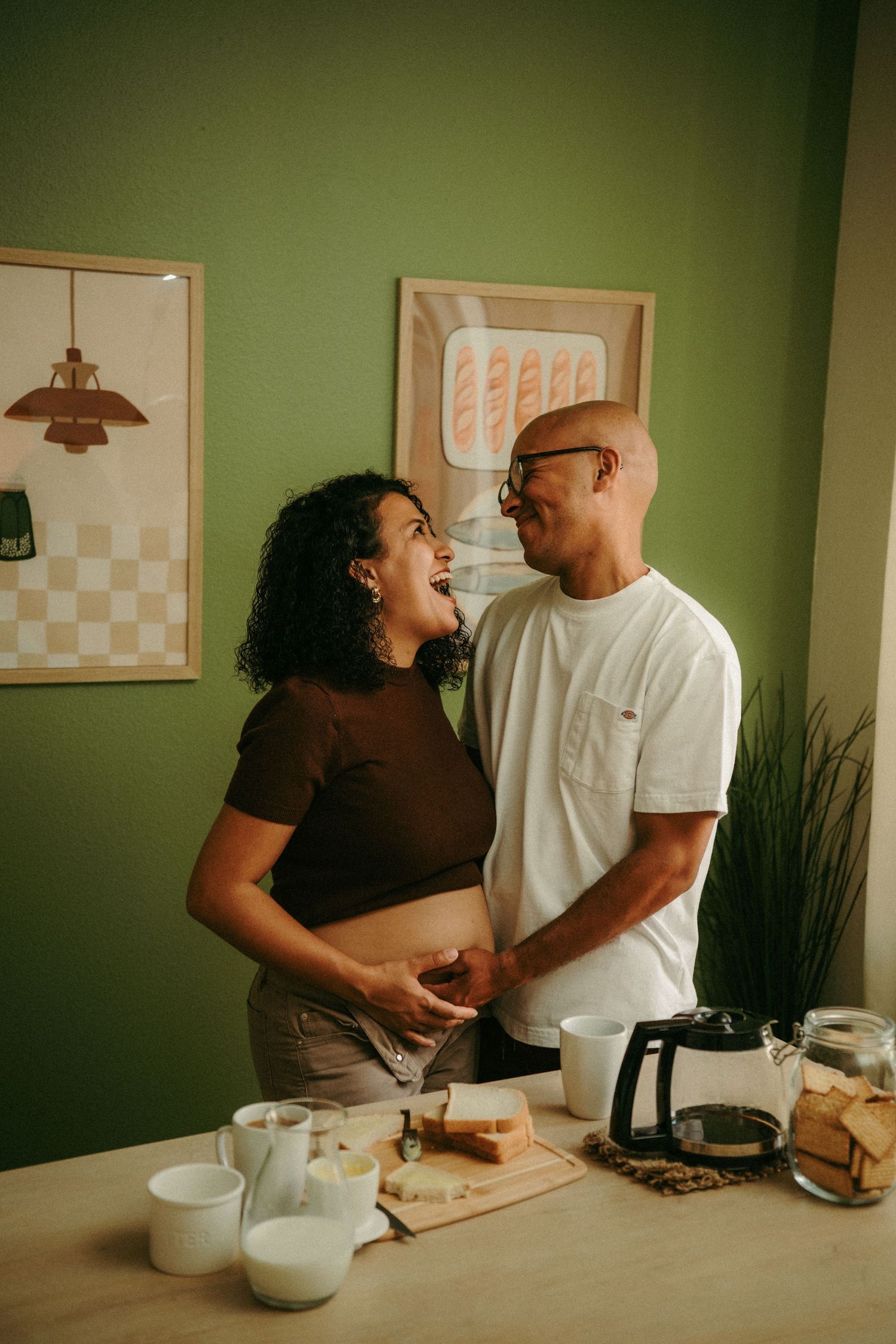 Happy couple sharing a laugh in a cozy green kitchen with coffee and breakfast items on the counter.