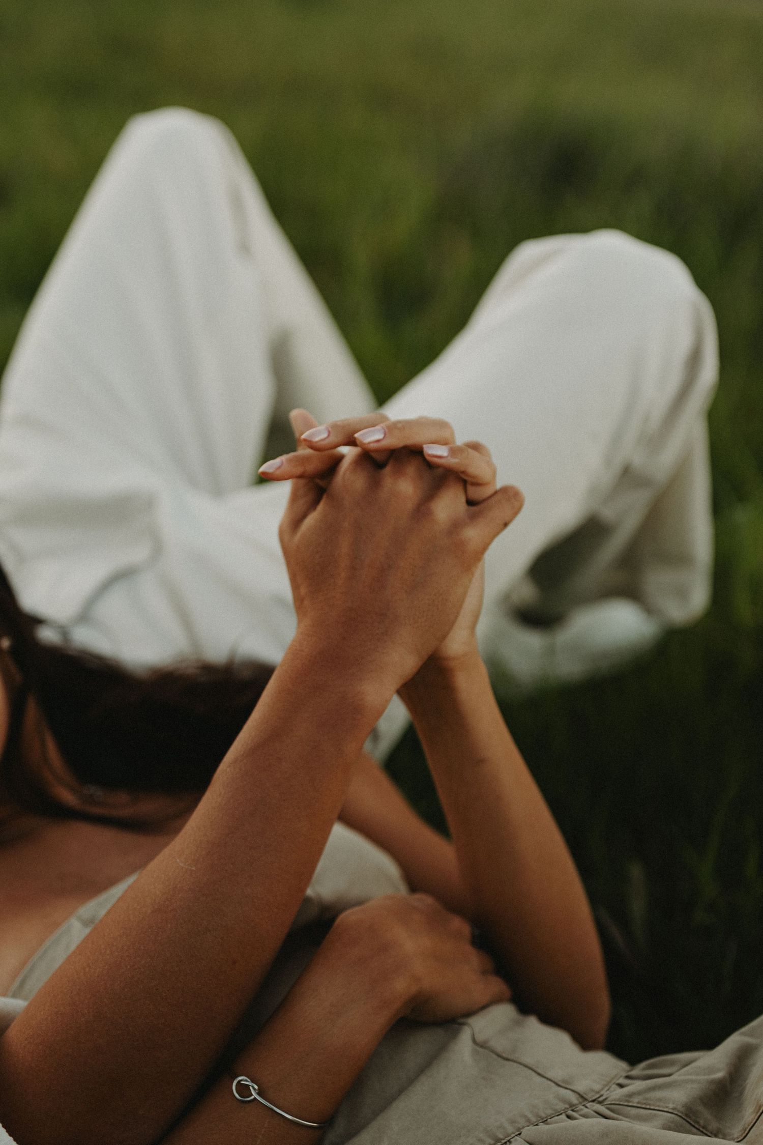 Two people lying in green grass holding hands together, one wearing white pants and a silver bracelet.