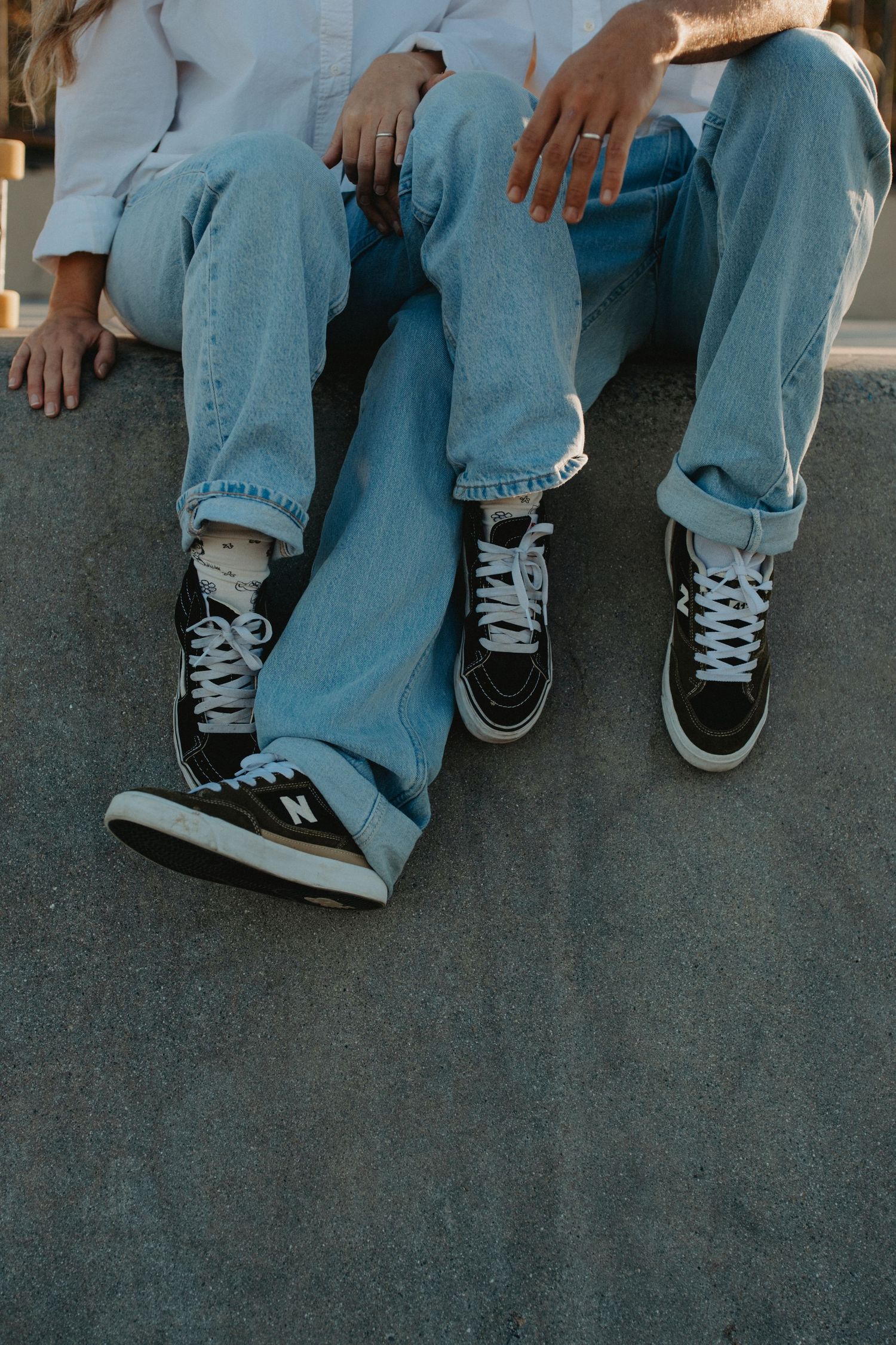 Couple sitting together wearing matching blue jeans and black high-top New Balance sneakers on concrete.