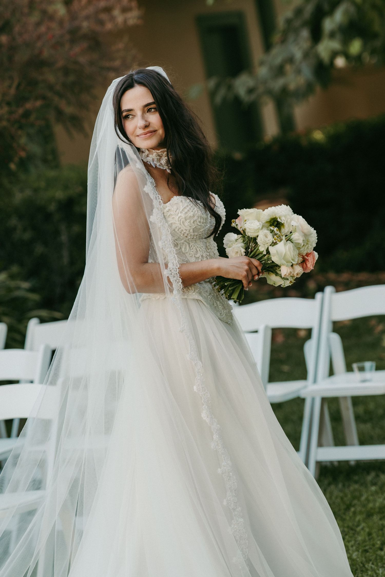 Bride in lace ball gown with veil holds white bouquet outdoors near white chairs.