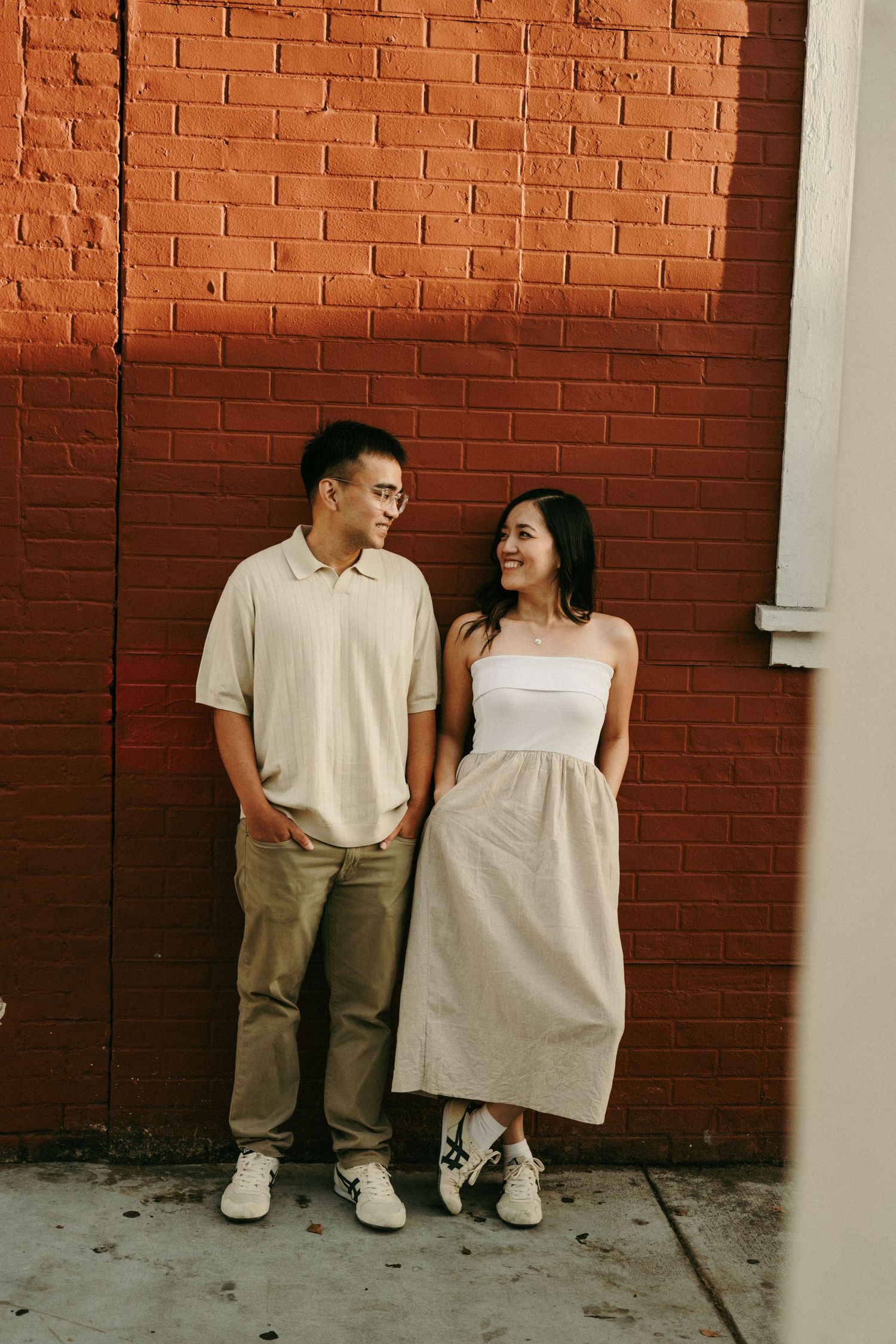 A couple smiles at each other leaning against a red brick wall, dressed in casual neutral tones.