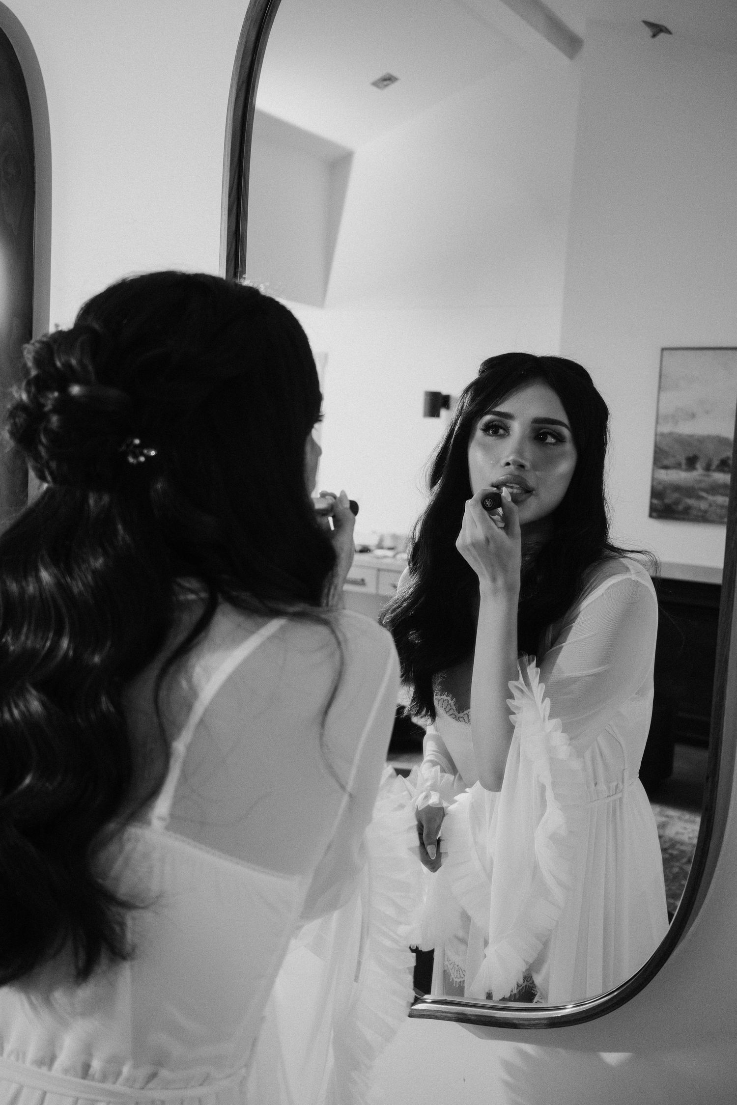 Black and white photo of dark-haired bride in white robe applying lipstick while looking in arched mirror.