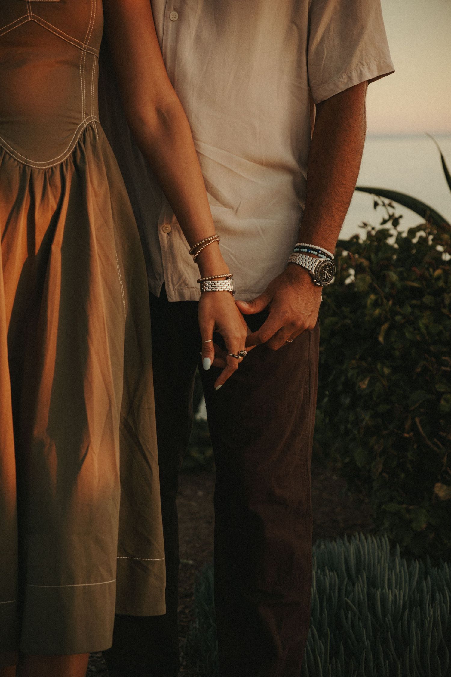Couple holding hands at sunset, wearing bracelets and rings, standing near coastal greenery.