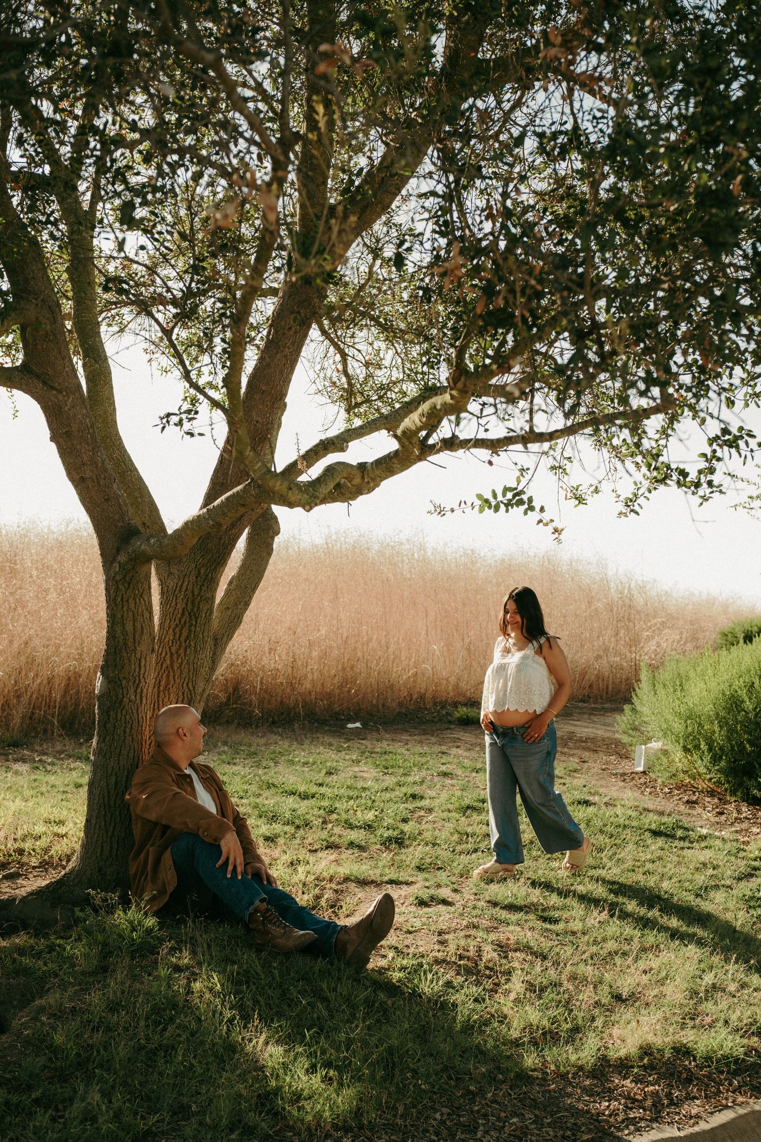 Man sitting against a tree while a woman stands nearby in a sunlit park with golden grass in the background.