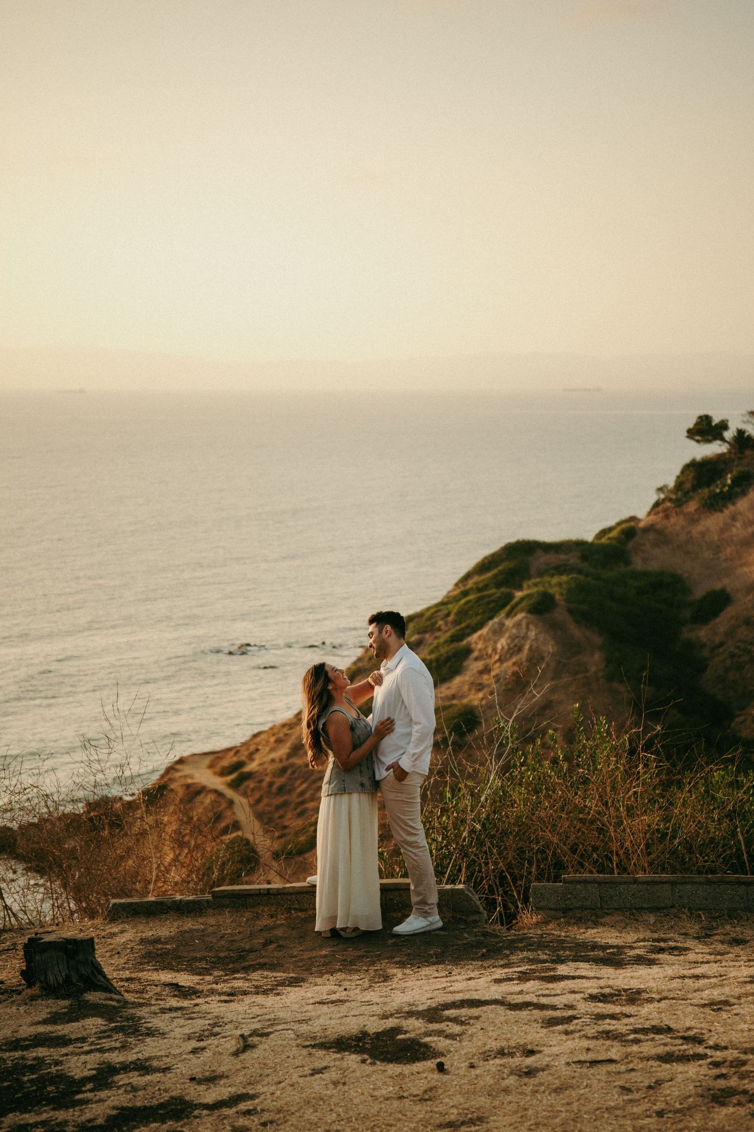 Couple embracing on a coastal cliff at sunset with ocean and golden hills in the background.