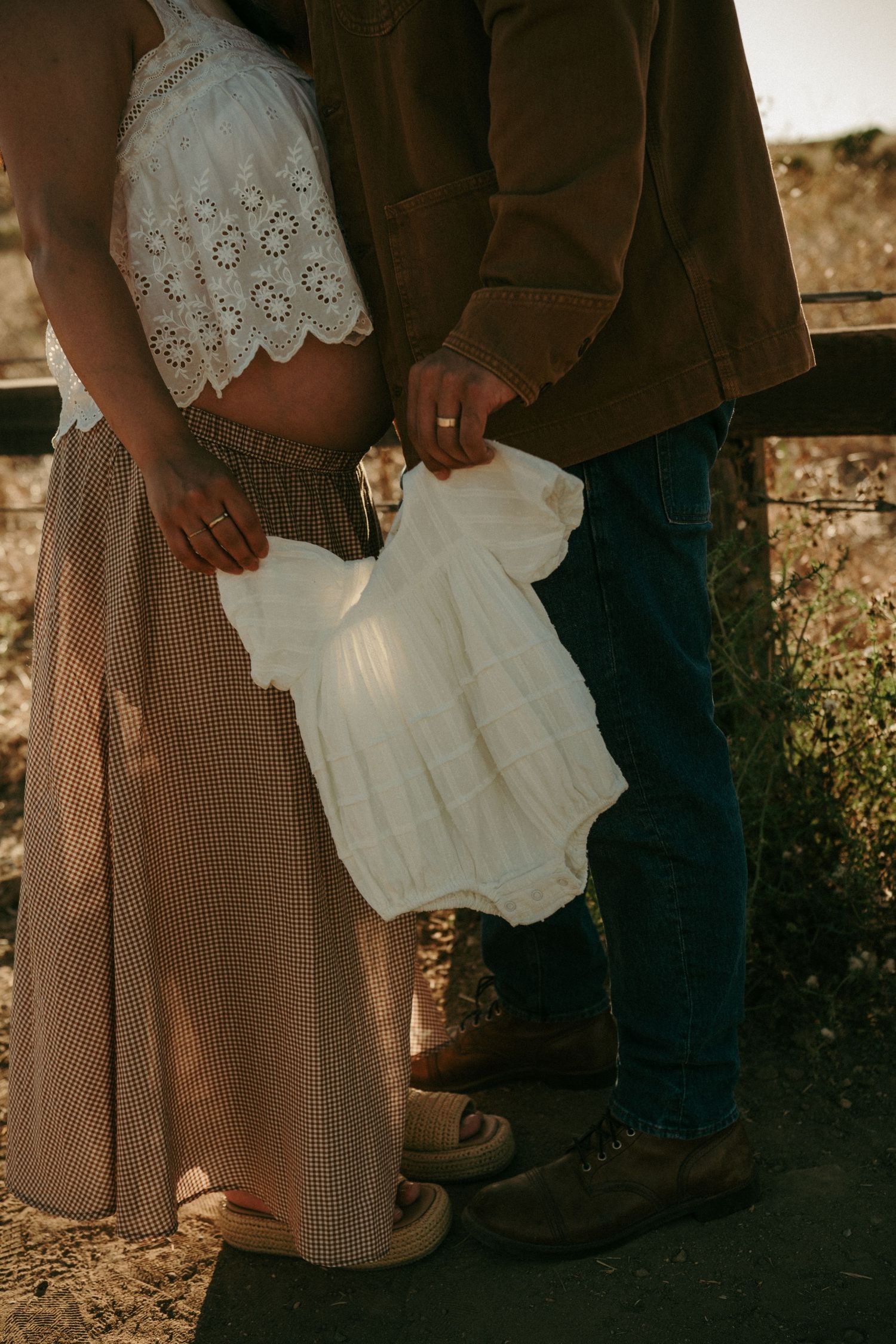 Pregnant couple holding a white baby onesie outdoors, woman in lace crop top and plaid skirt, man in brown jacket and jeans.