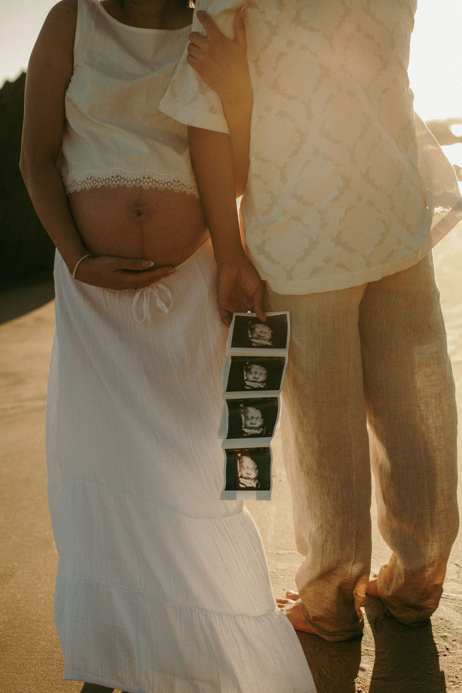 Pregnant couple standing outdoors at sunset, woman in white dress holding baby bump, man holding ultrasound photos.