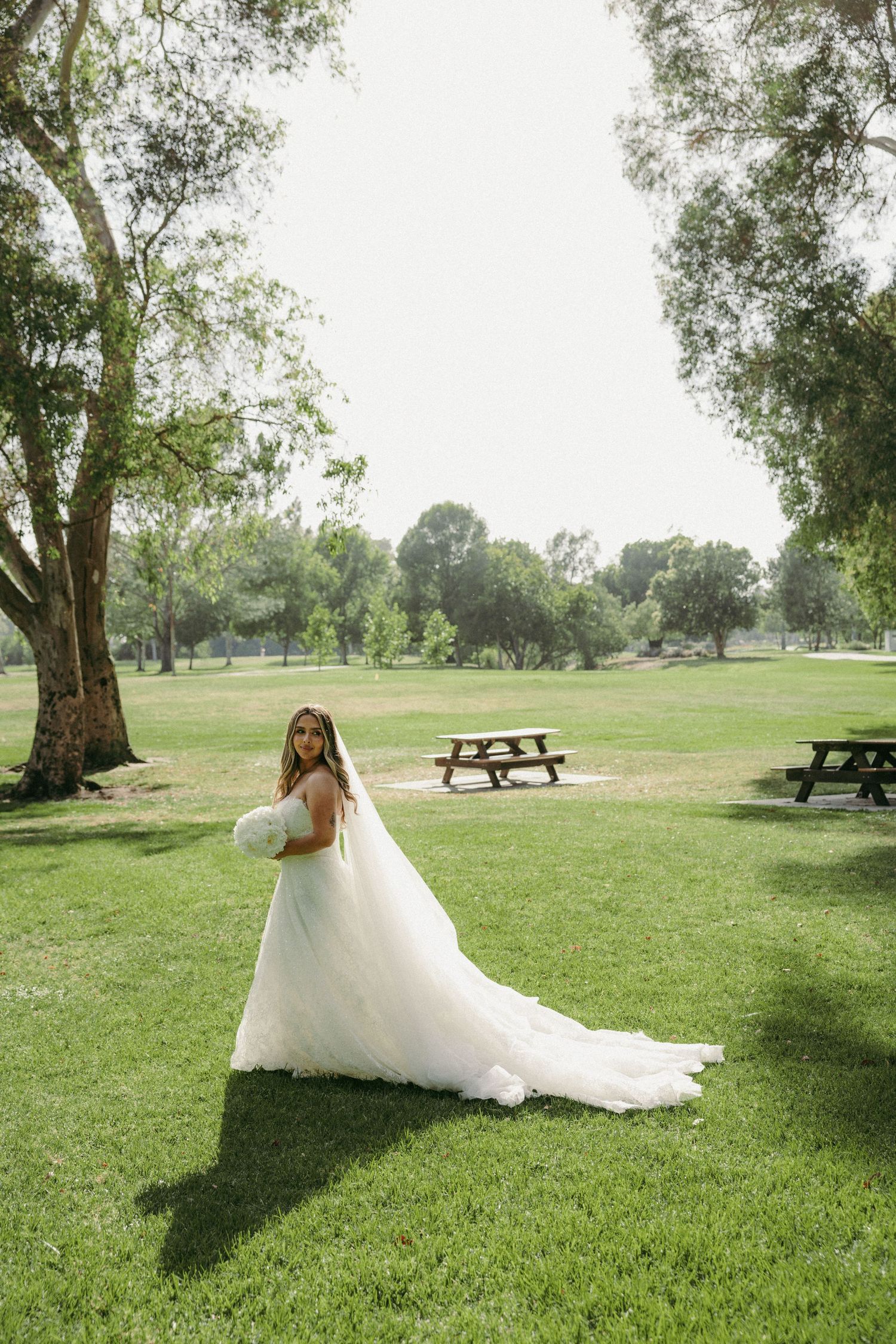 Bride in white gown with long train stands on lush green lawn surrounded by trees during outdoor bridal portrait session.