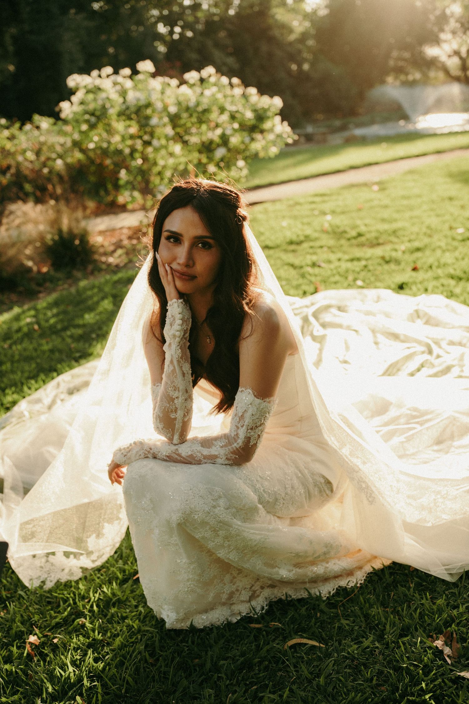 Bride in lace wedding gown and gloves sits gracefully on green grass in a sunlit garden setting.